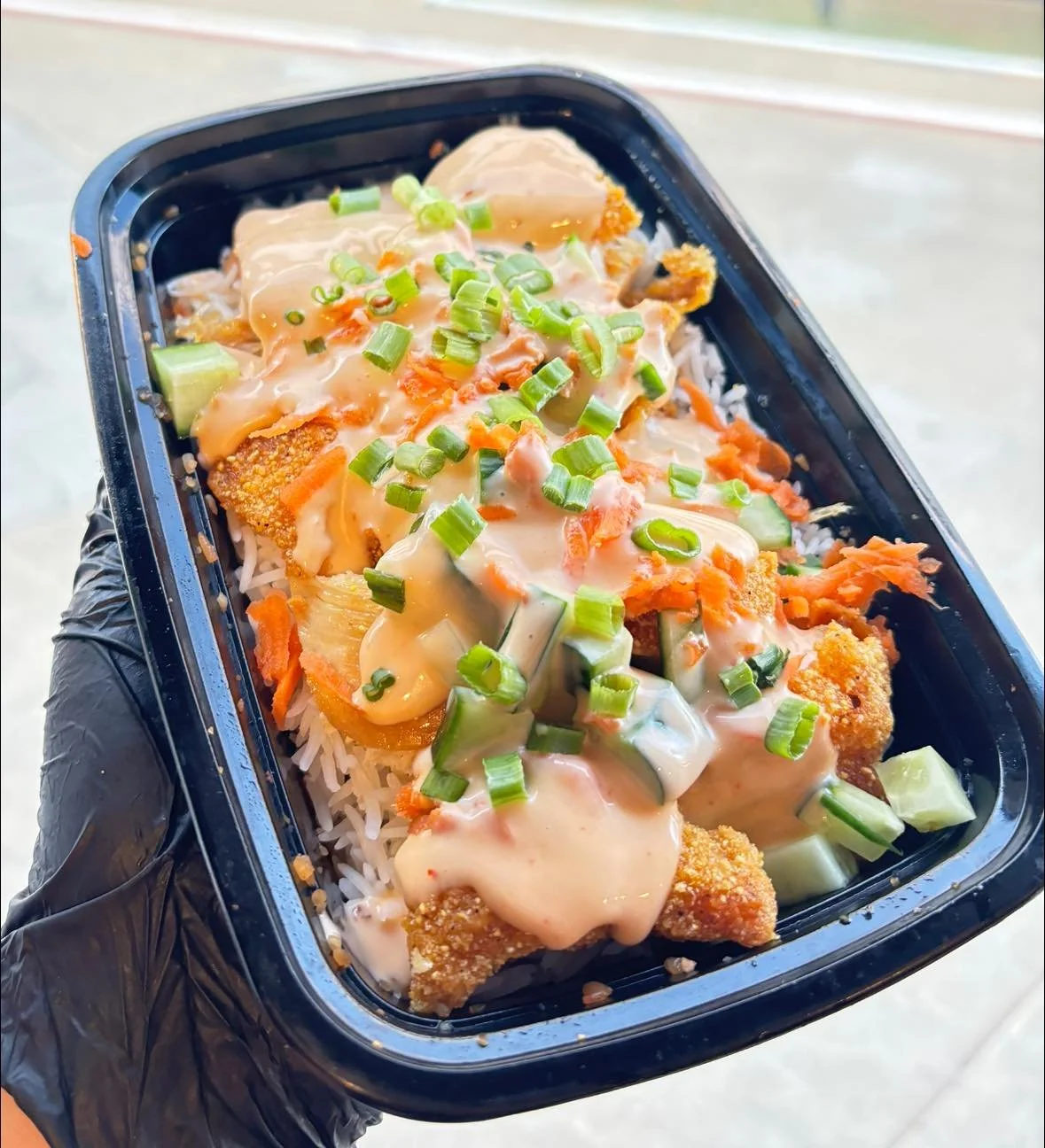 Takeout black container of rice topped with fried fish, chopped green onions, and creamy sauce with cucumbers.