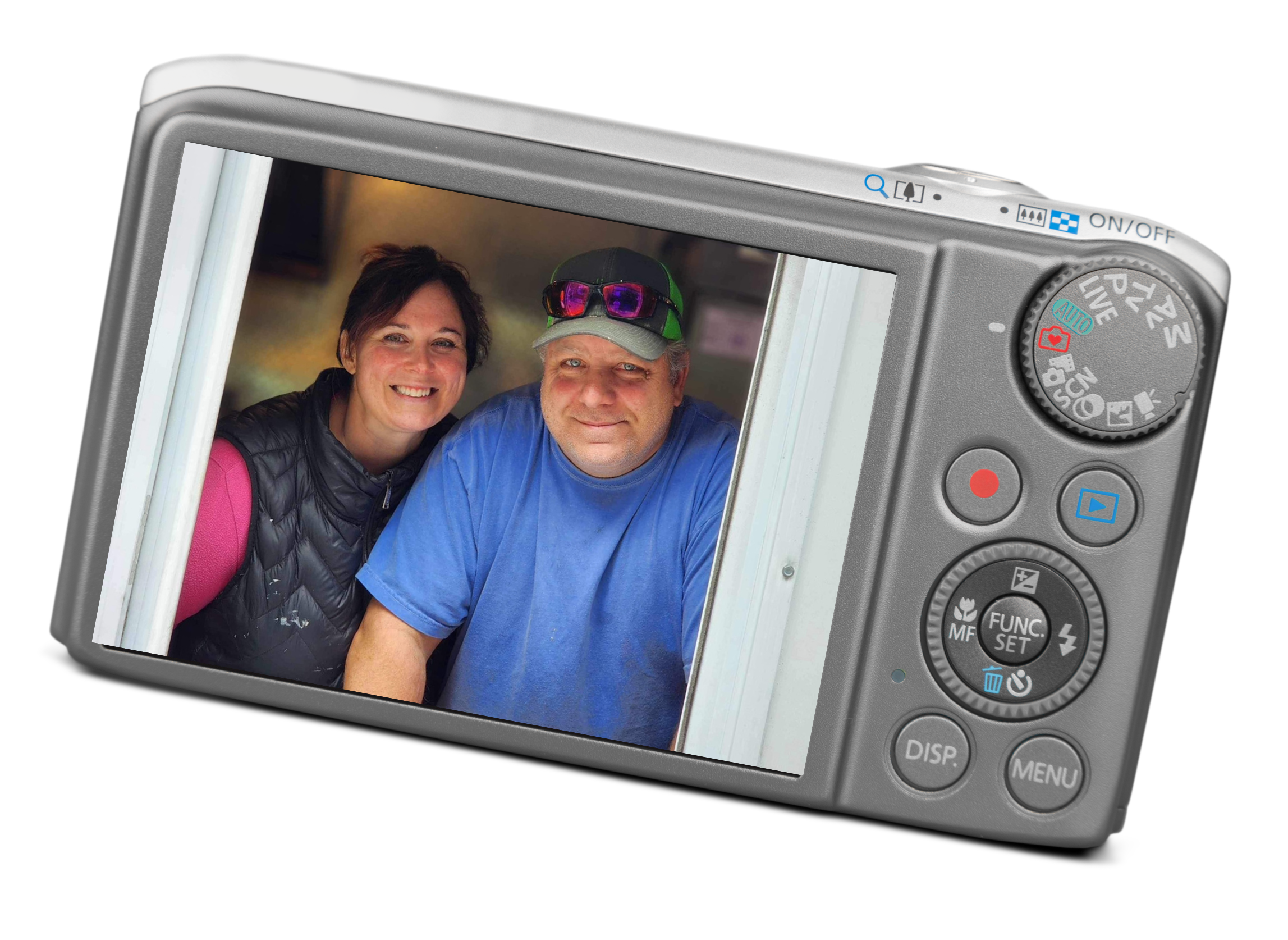 A digital camera displaying a photo of a woman and a man smiling, both indoors, with the woman wearing a black vest and pink shirt, and the man in a blue shirt and baseball cap with sunglasses on top.