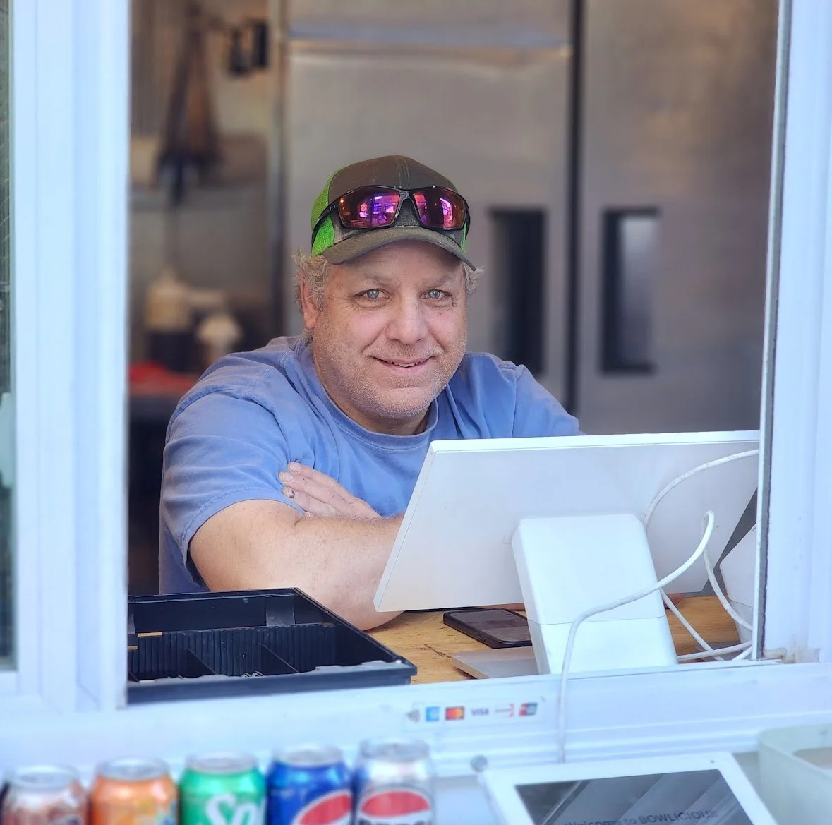 A man with gray hair and blue eyes, wearing a gray cap with sunglasses on top, sitting at a food stand window and smiling at the camera.