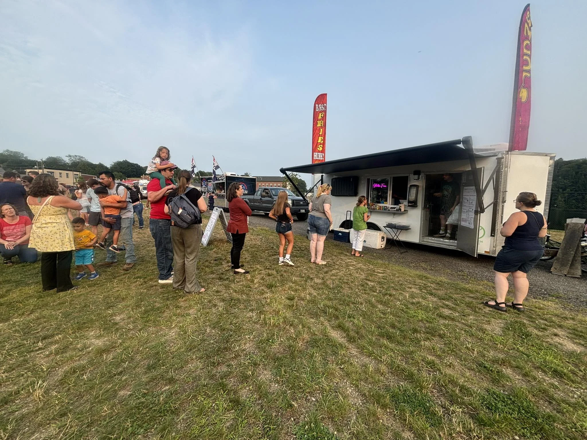People waiting in line at a food truck during an outdoor event, with a mix of adults and children on a grassy area.