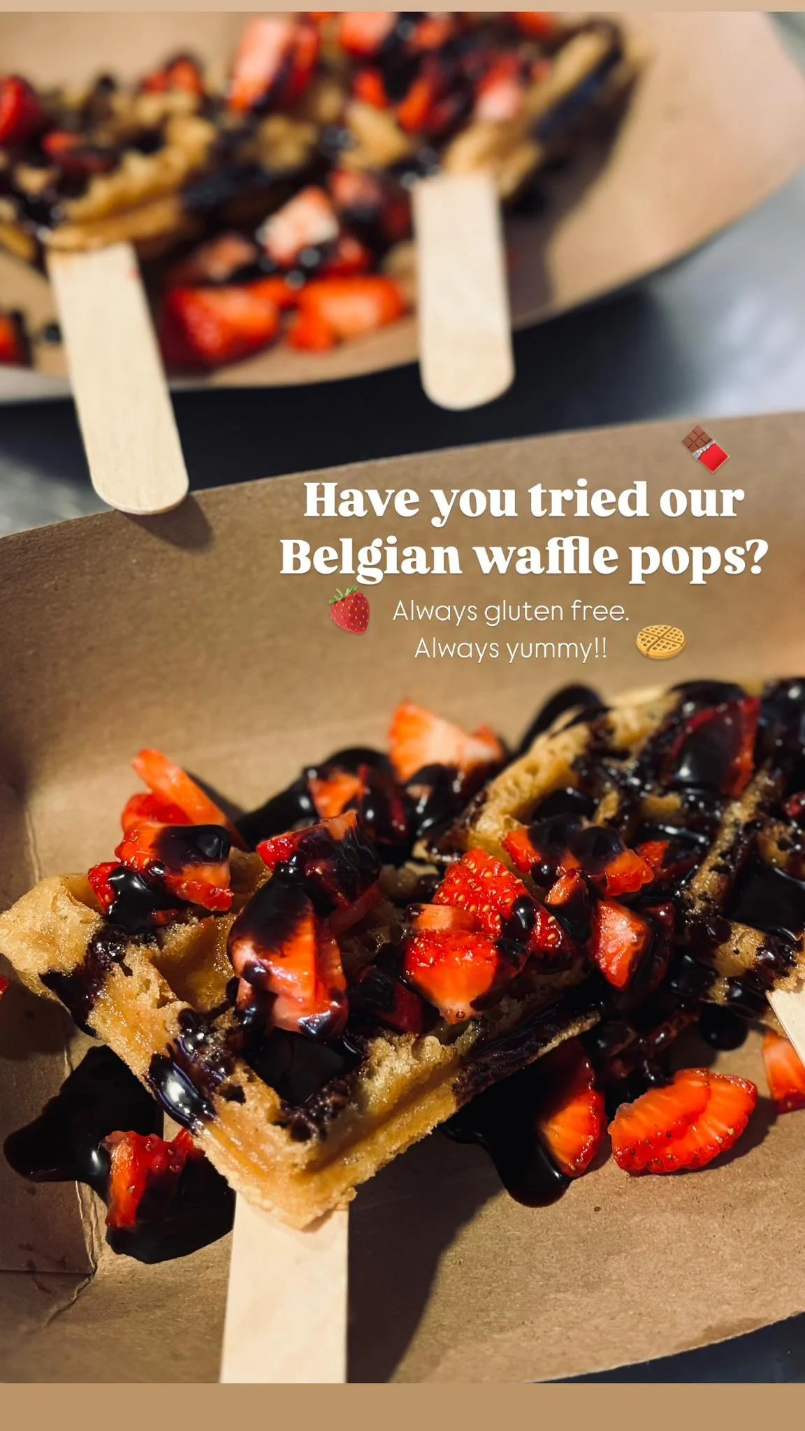 Belgian waffle topped with strawberries and drizzled with chocolate sauce, served on a paper-lined tray with a wooden stick.