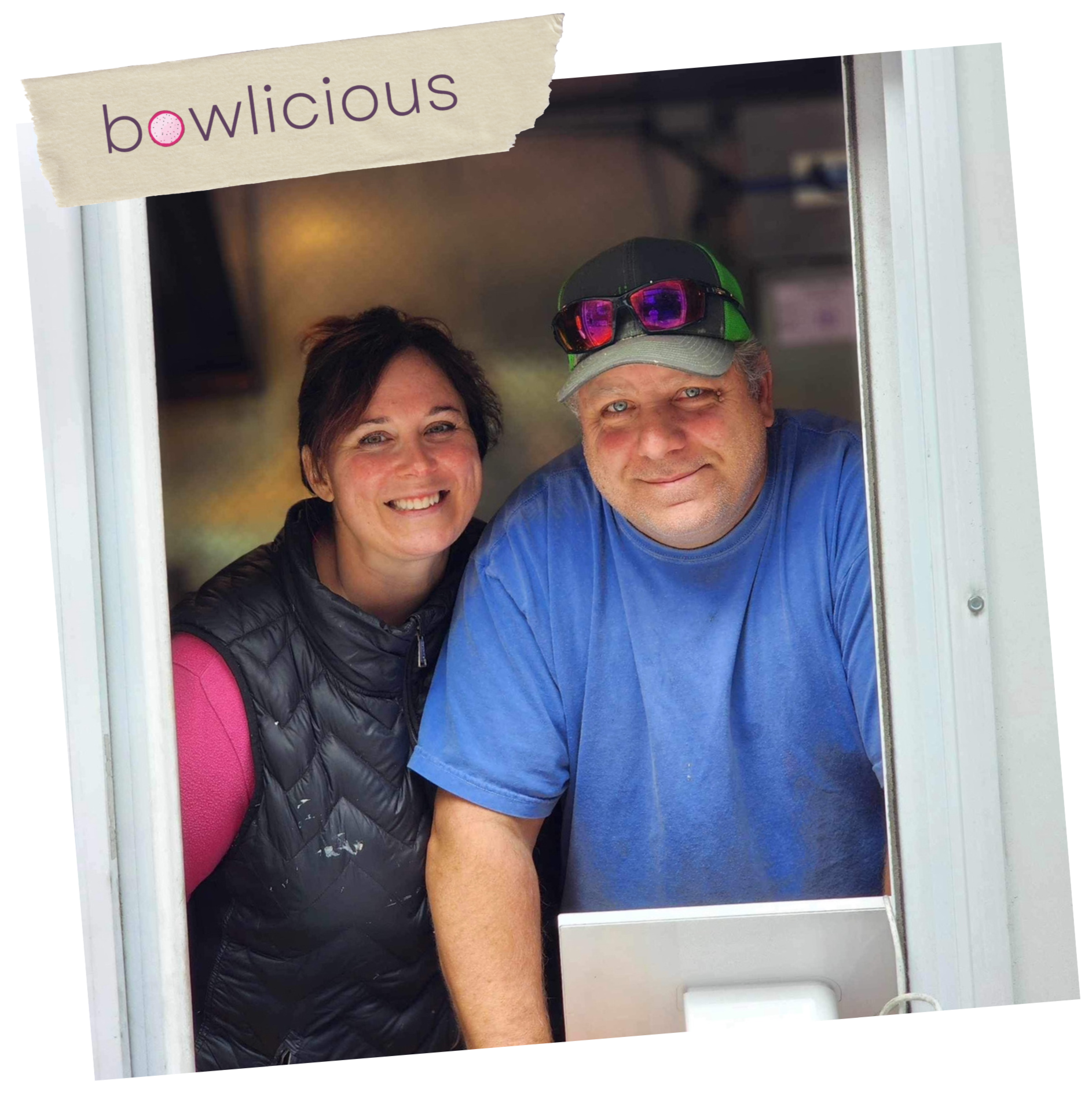 A woman and a man smiling at a food truck window. The woman has short dark hair and is wearing a black puffy vest and pink long sleeve shirt. The man has a baseball cap with sunglasses on it, blue shirt, and is slightly tanned.