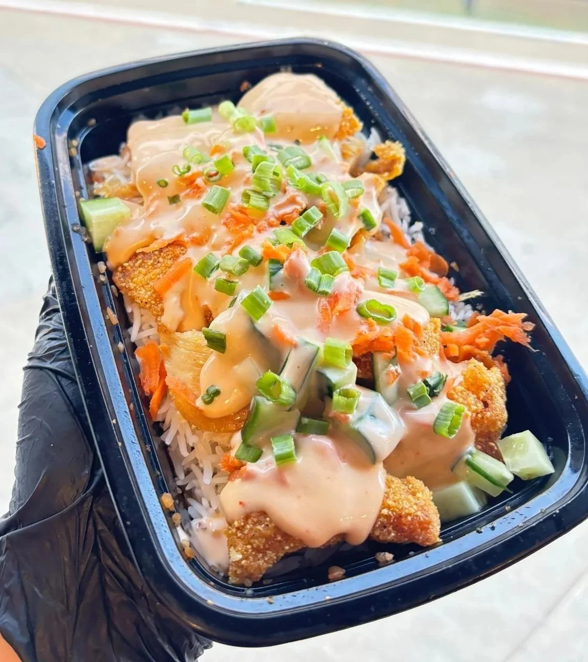 Takeout container of fried fish fillets topped with creamy sauce, chopped green onions, and vegetables over rice.