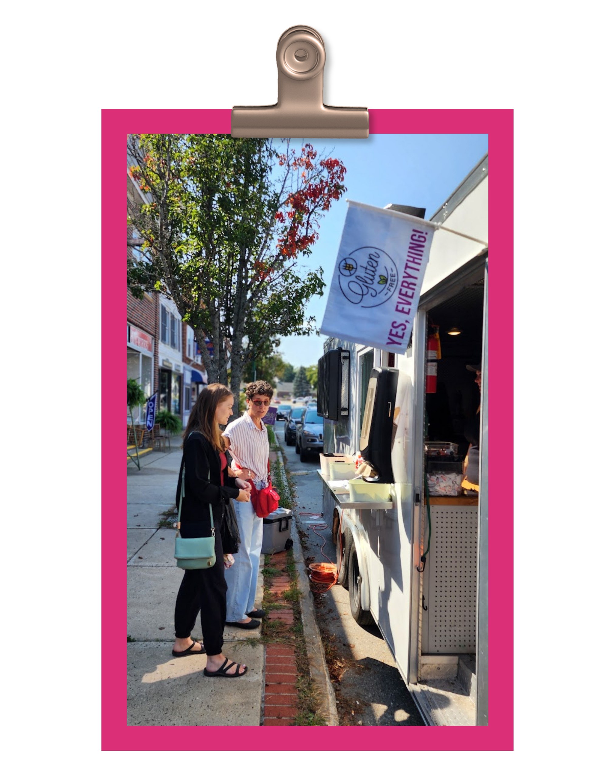 Two women stand in line at a food truck on a sunny street, with trees and shops visible in the background. The food truck has a banner that reads 'YES, EVERYTHING!'