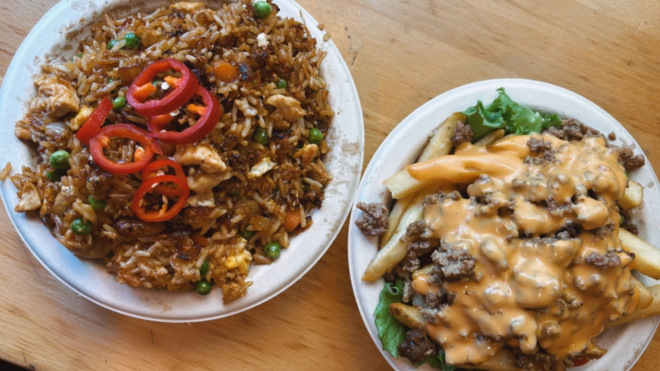 Two plates of fast food on a wooden table. The left plate contains fried rice with peas, diced vegetables, pieces of chicken, and sliced red chilies on top. The right plate has French fries topped with ground beef, shredded cheese, and a drizzle of cheese sauce, with some lettuce underneath.