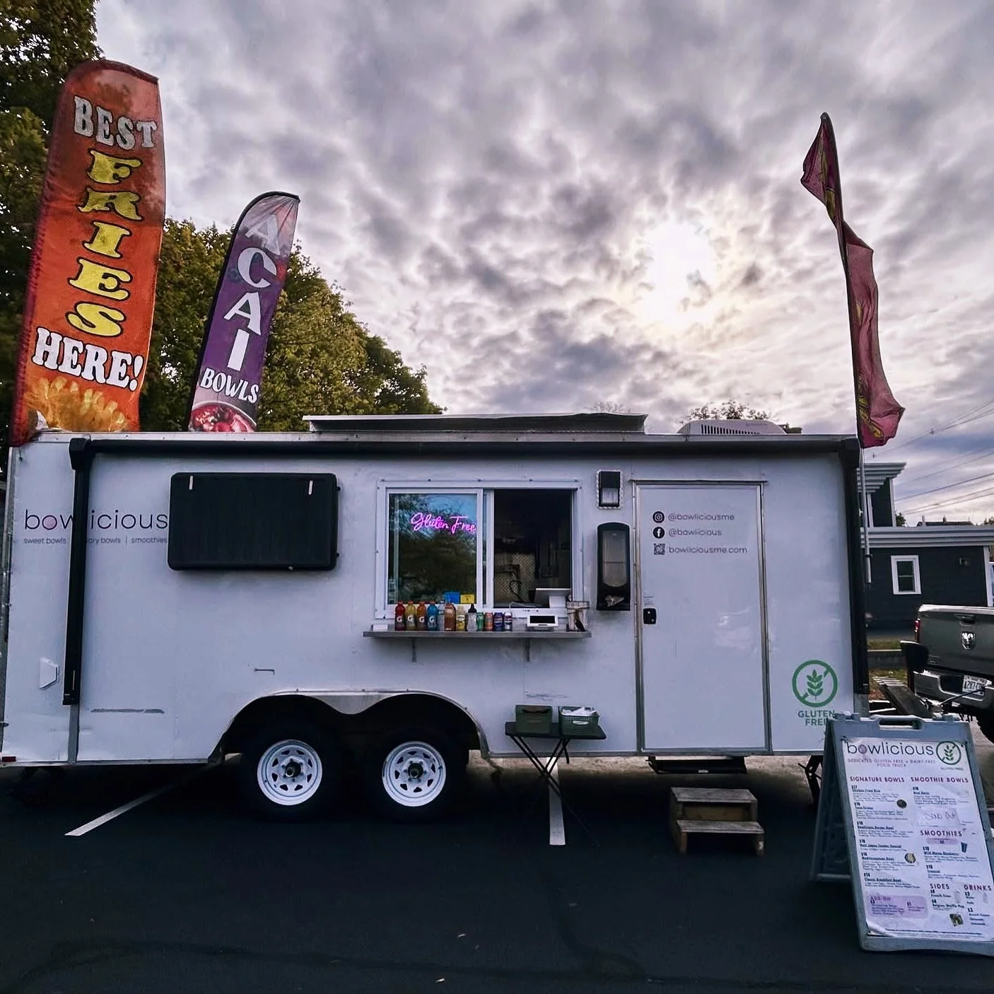A white food truck named 'Bowlicious' parked in a lot on a cloudy day. The truck advertises gluten-free options and has colorful bottles on a small shelf outside. There are flags and a menu board nearby, with a neon sign that reads 'Gluten Free' in the window.