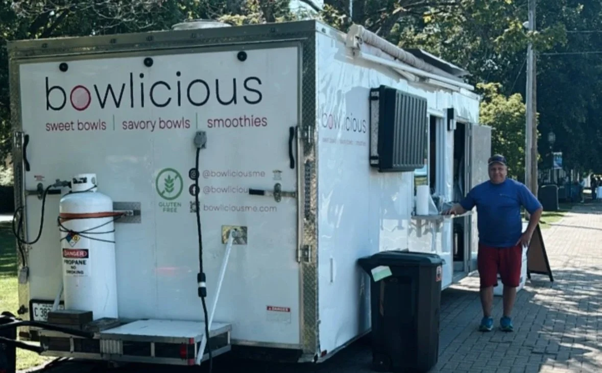 A food truck named 'bowlicious' serving sweet bowls, savory bowls, and smoothies, with a person standing next to it on a sidewalk.