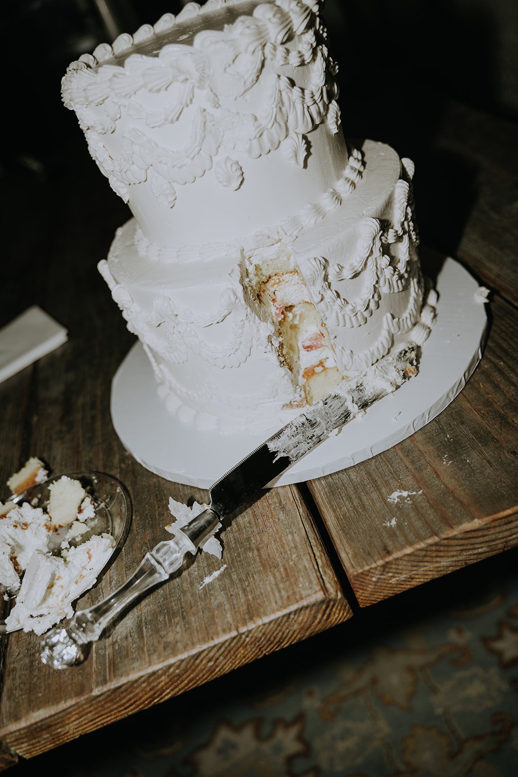 White wedding cake with decorative icing, partially cut with a serrated knife on a wooden table, with a glass dish of cake scraps nearby.