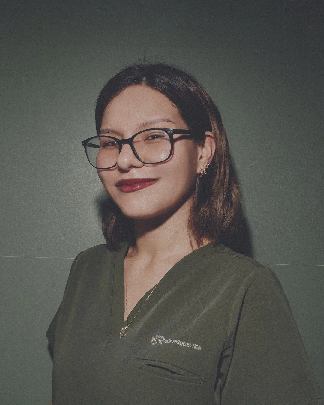 A woman with shoulder-length brown hair, wearing glasses and a dark green shirt, smiling against a plain background.