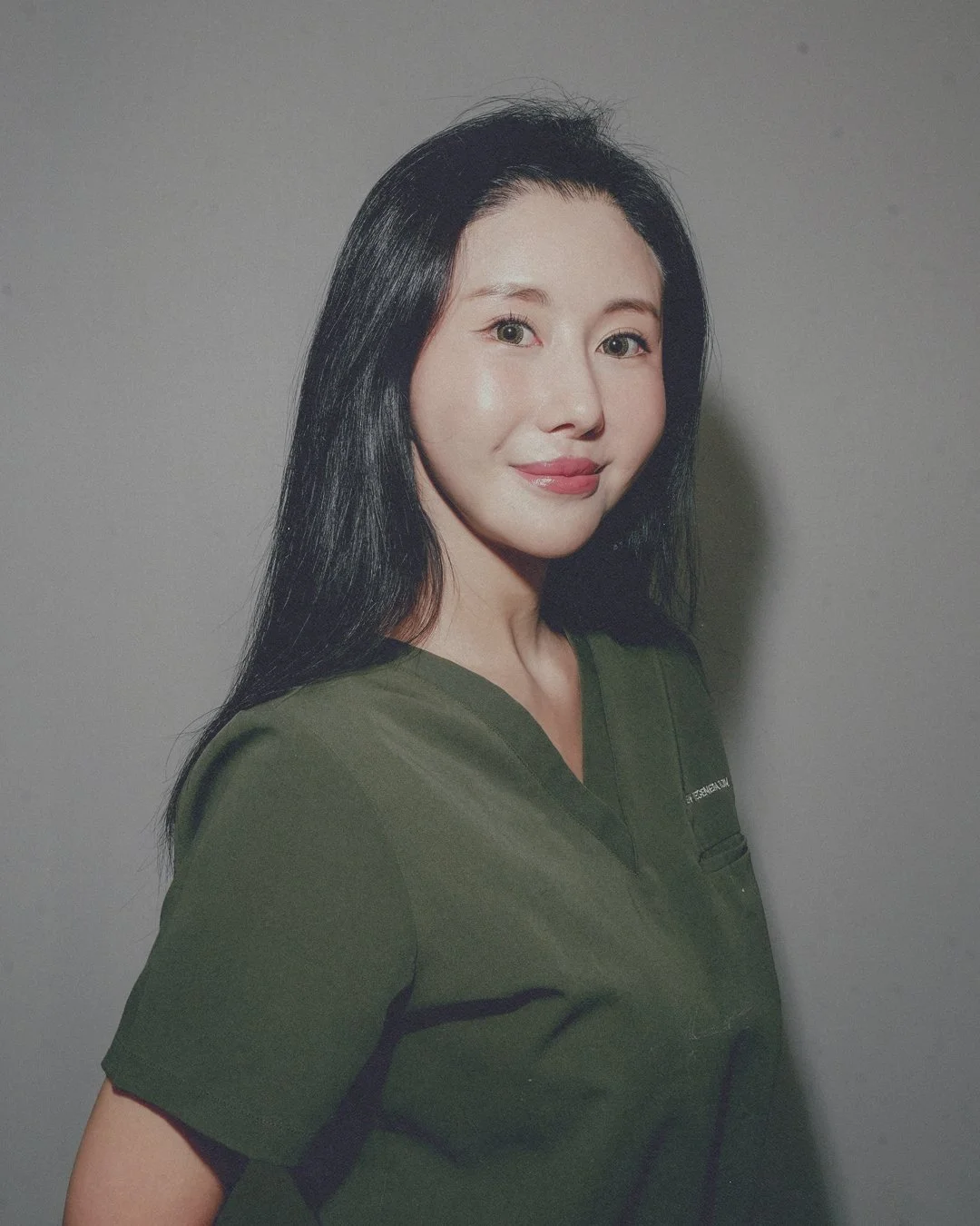 Portrait of a woman with long black hair, wearing a dark green V-neck top, smiling softly against a plain gray background.