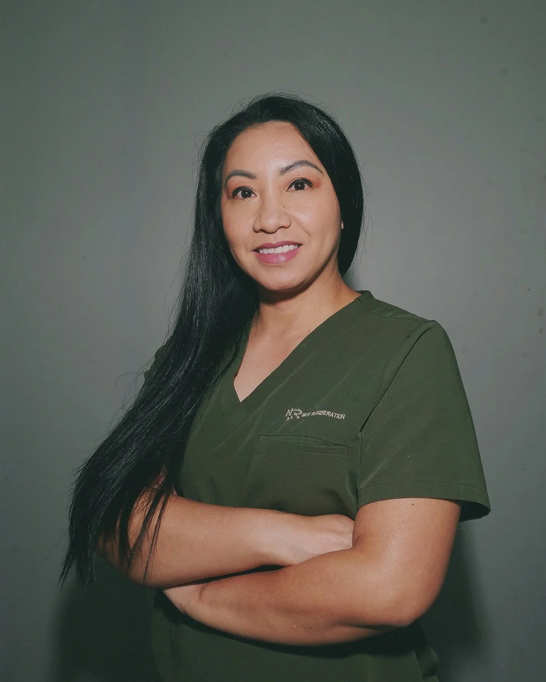 A woman with long black hair, wearing a dark green scrub top, standing with arms crossed against a gray background.