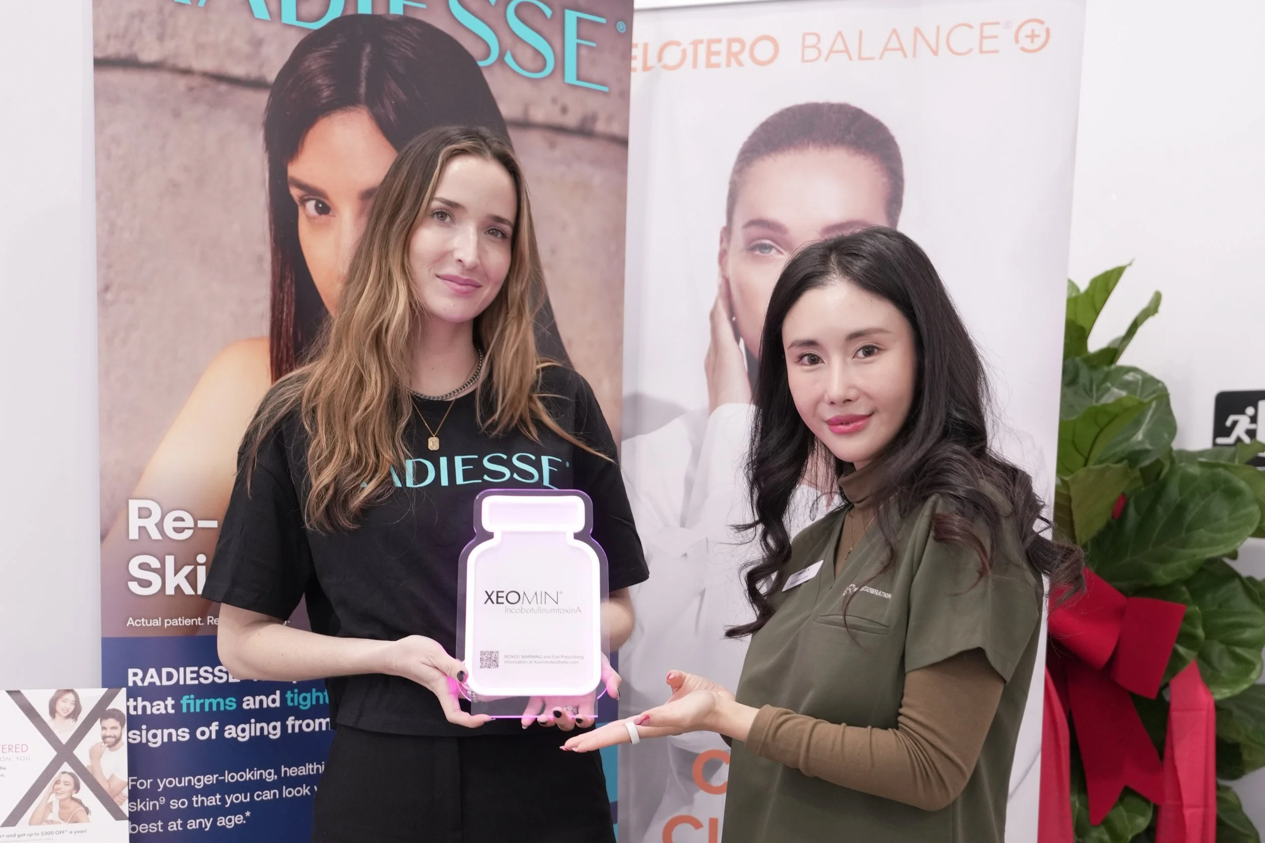Two women at a promotional event for skincare or anti-aging products; one woman with long brown hair is holding a large model of a supplement bottle labeled XEO MIN, and the other woman with long dark hair is supporting the bottle's base. Behind them are promotional banners featuring images of women and product information.