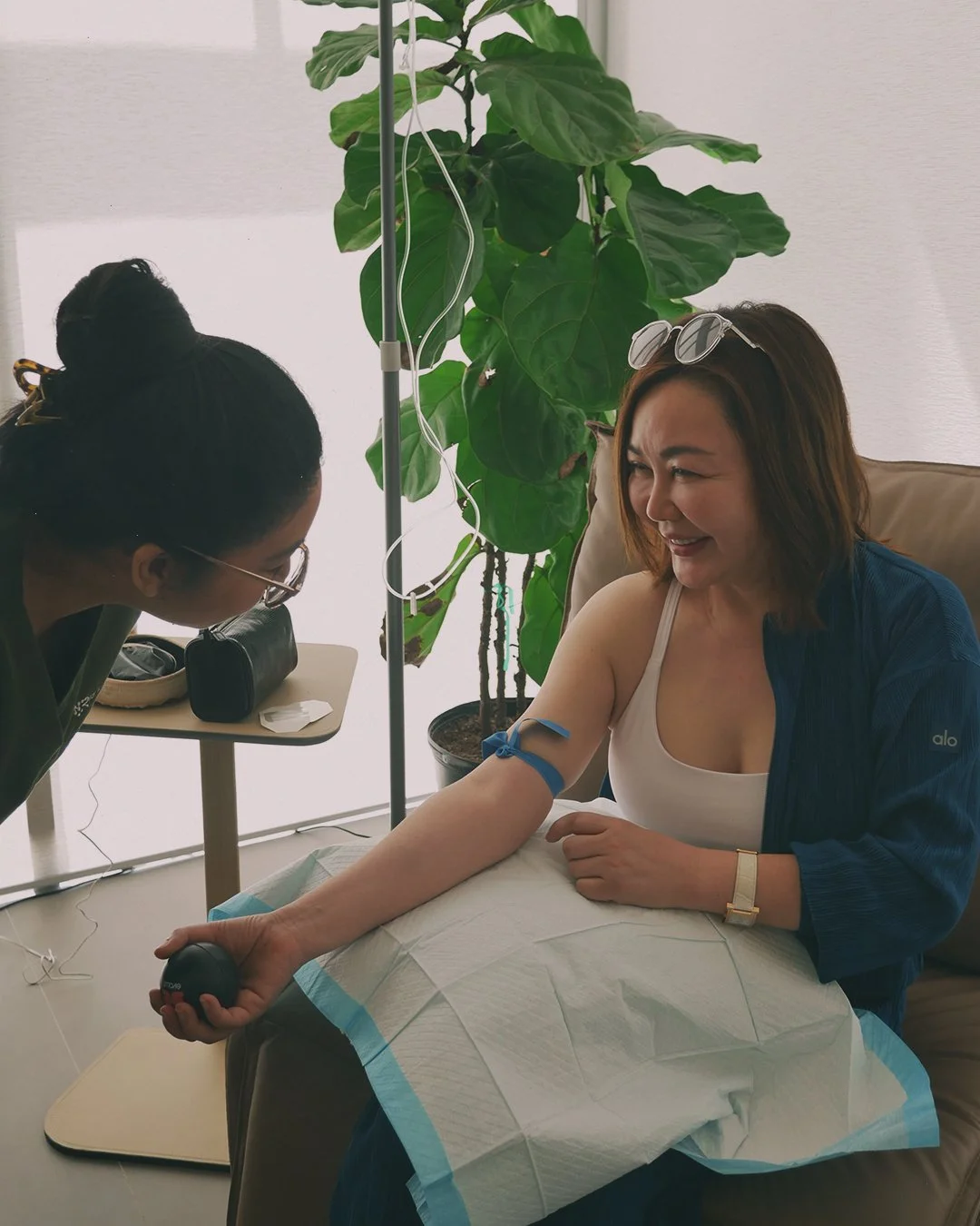 A woman sitting in a medical chair, smiling and looking at a nurse, while the nurse tests her blood sugar using a glucometer. The woman has a tourniquet on her arm, and there is a large green plant in the background.
