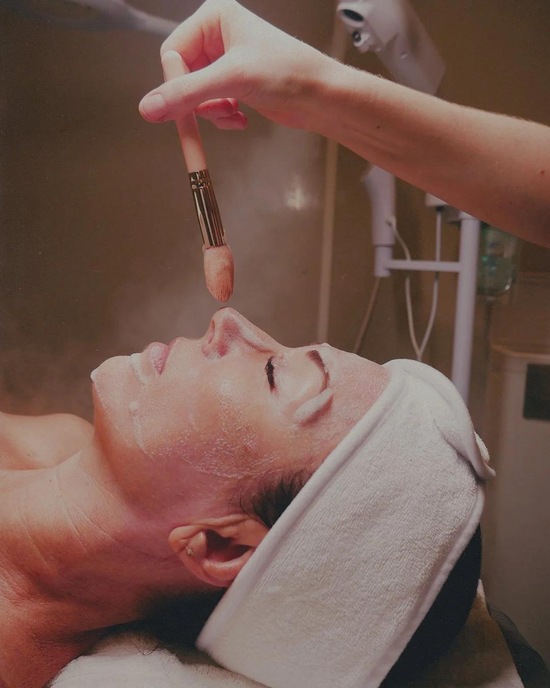 A person lying down with a headband, receiving a facial treatment. A beautician applies a substance to the person's face with a brush.