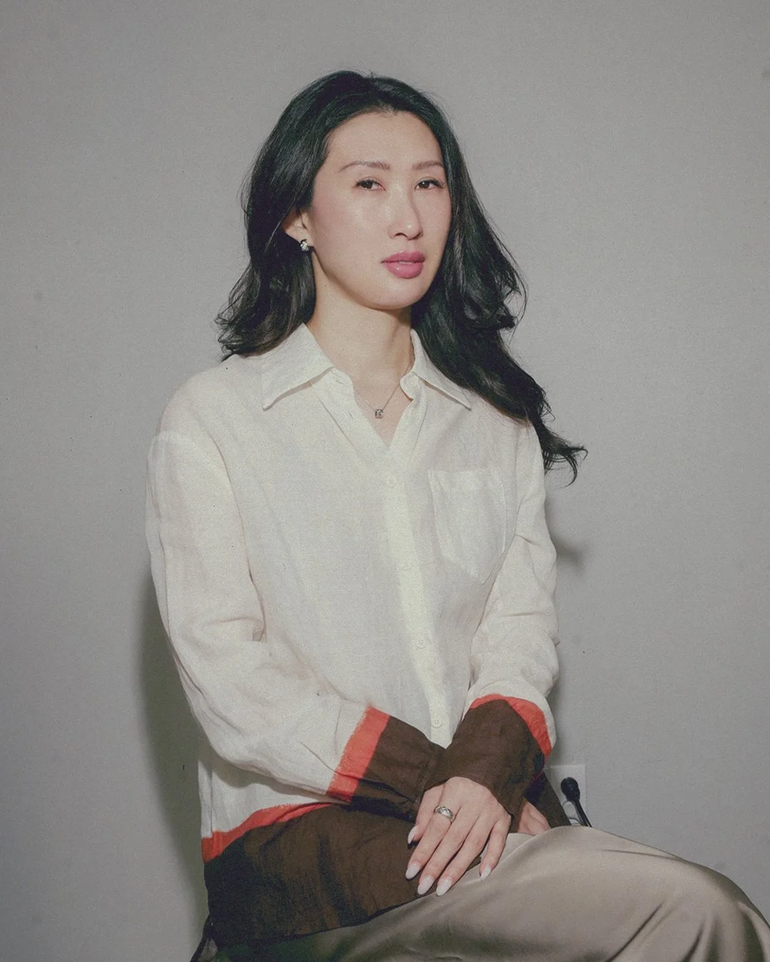 A woman with long black hair and light skin, wearing a white shirt with brown and orange accents on the sleeves, sitting against a plain gray background.