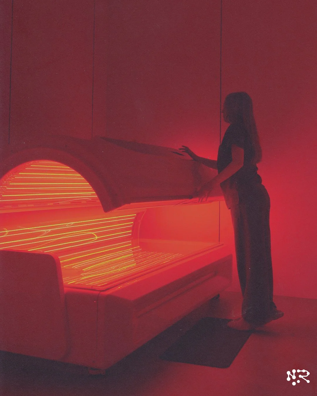Person standing next to a red tanning bed in a dimly lit room.