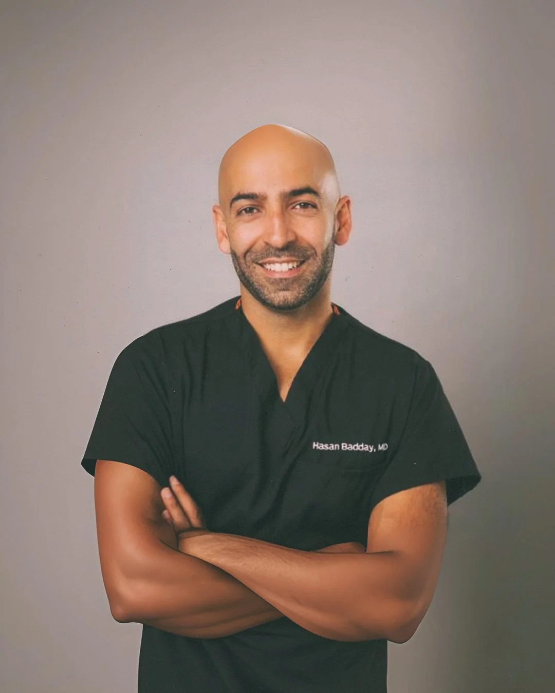 A smiling male doctor in a black medical uniform with his arms crossed, standing against a plain background.