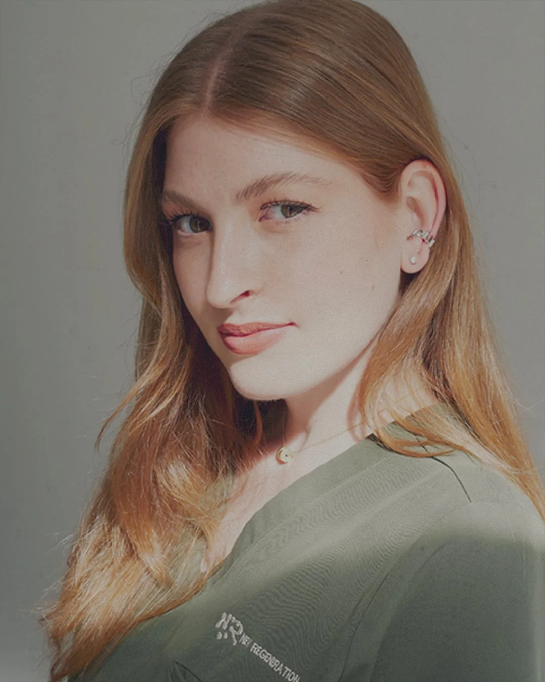 A woman with long, wavy red hair wearing a green shirt, earrings, and a necklace. She is looking at the camera with a slight smile.