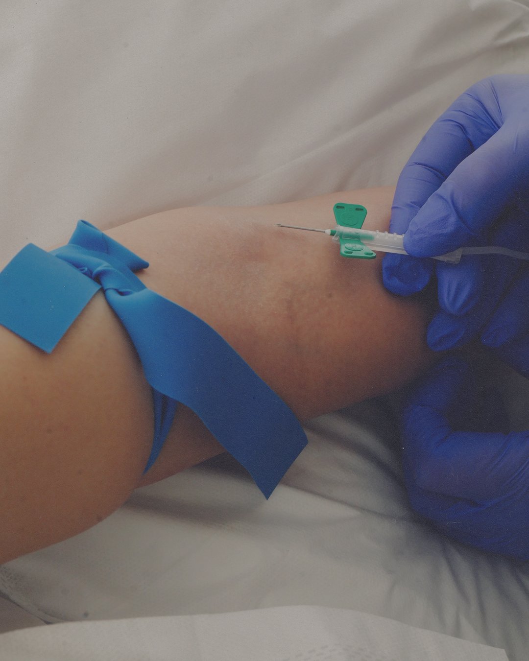 A person is getting a shot injected into their arm by a healthcare worker wearing blue gloves, with blue medical tape on the arm.