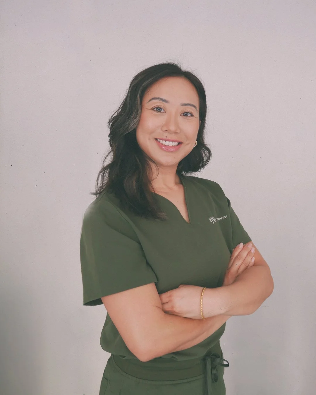 Smiling woman with dark wavy hair wearing green scrubs standing against a plain light-colored wall.