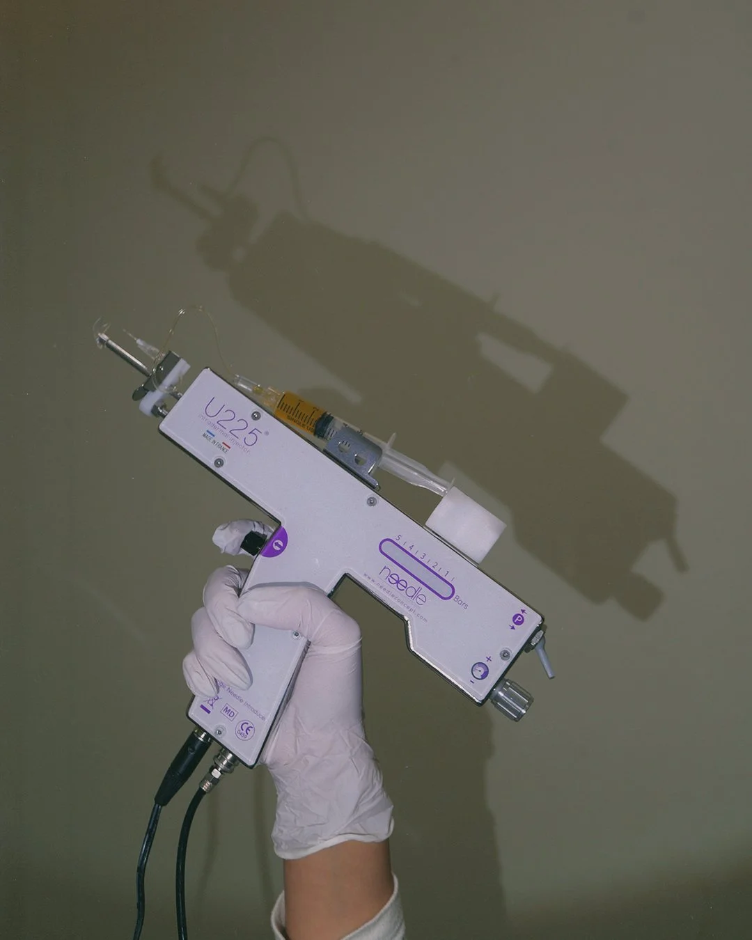 A person's hand wearing a white glove holding a medical device with a syringe attached, casting a shadow on a beige wall.