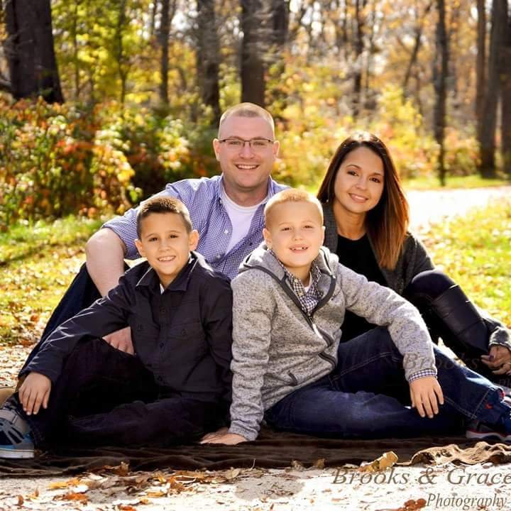 Family of four sitting outdoors in a park during fall, smiling, with colorful autumn leaves and trees in background.