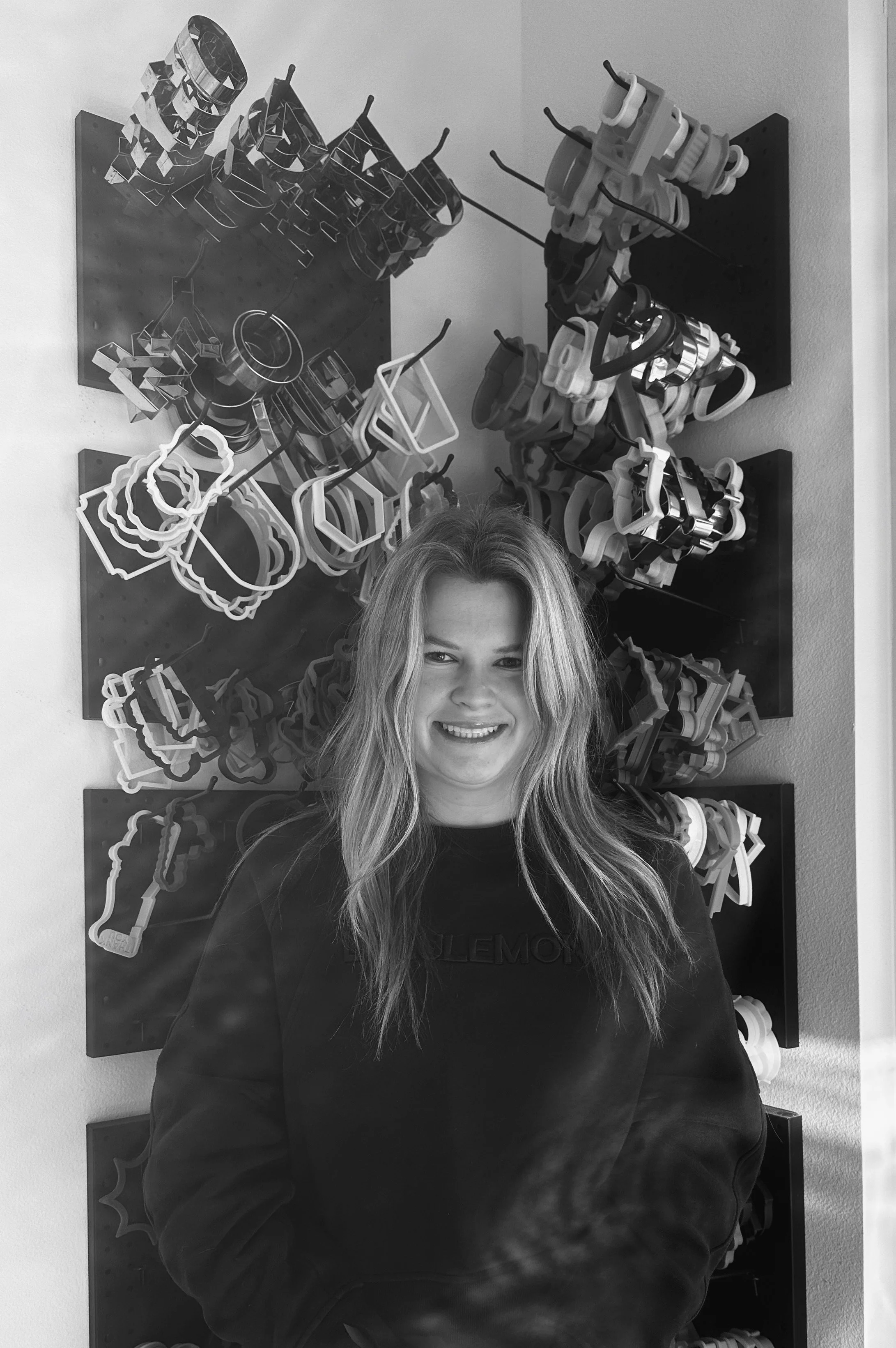 A young woman with long, wavy hair smiling in front of a wall display of cookie cutters.