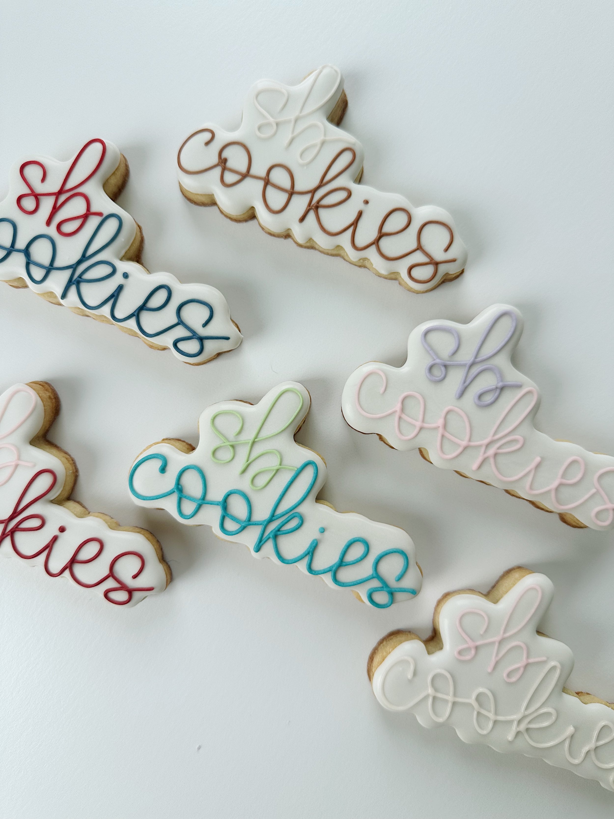 Decorated cookies with white icing and colorful cursive writing that spells out "Cookies" in different pastel colors, arranged on a white surface.