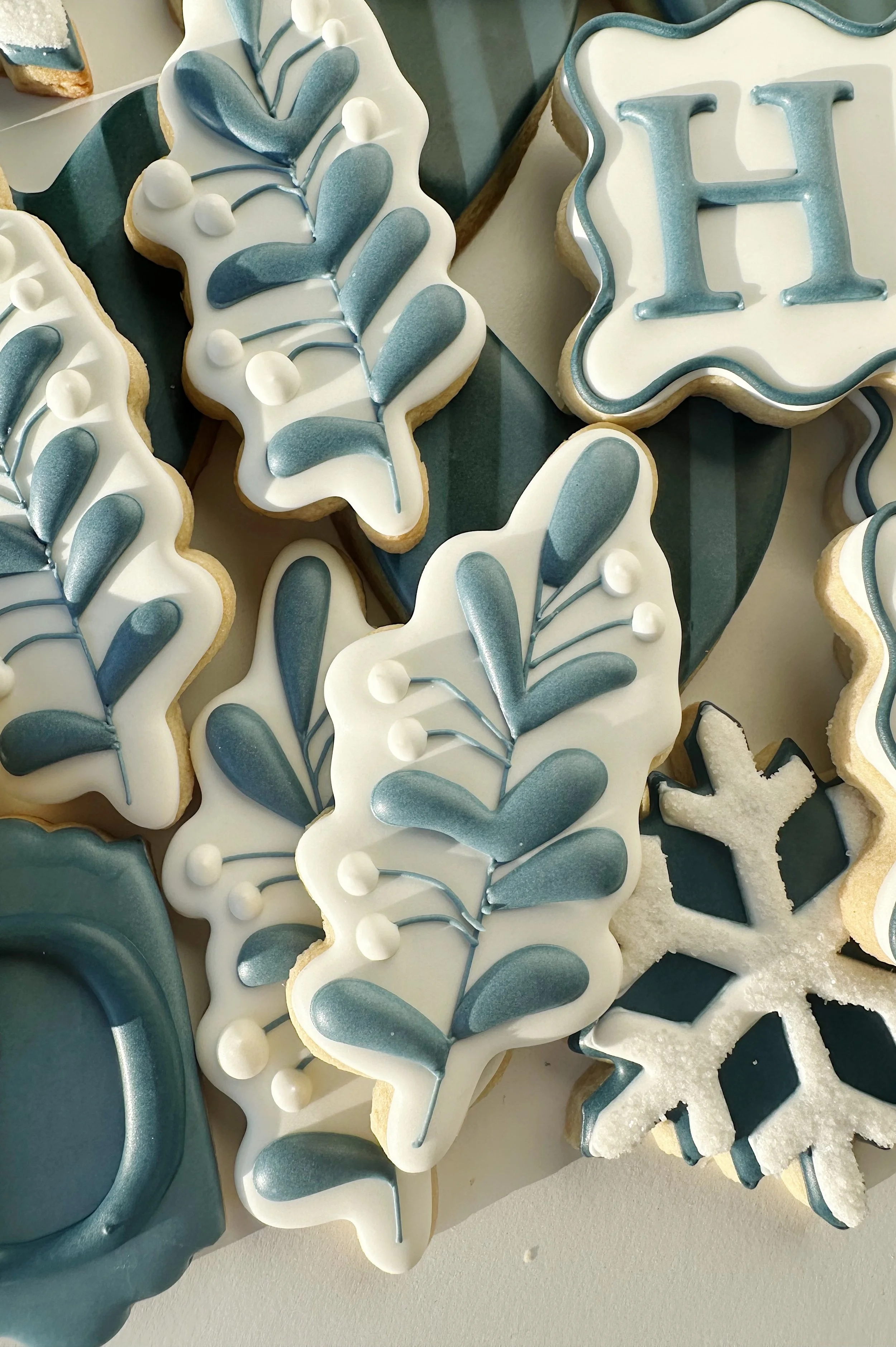 Decorated holiday cookies with blue, white, and silver icing, shaped like leaves, snowflakes, and a pill-shaped ornament.