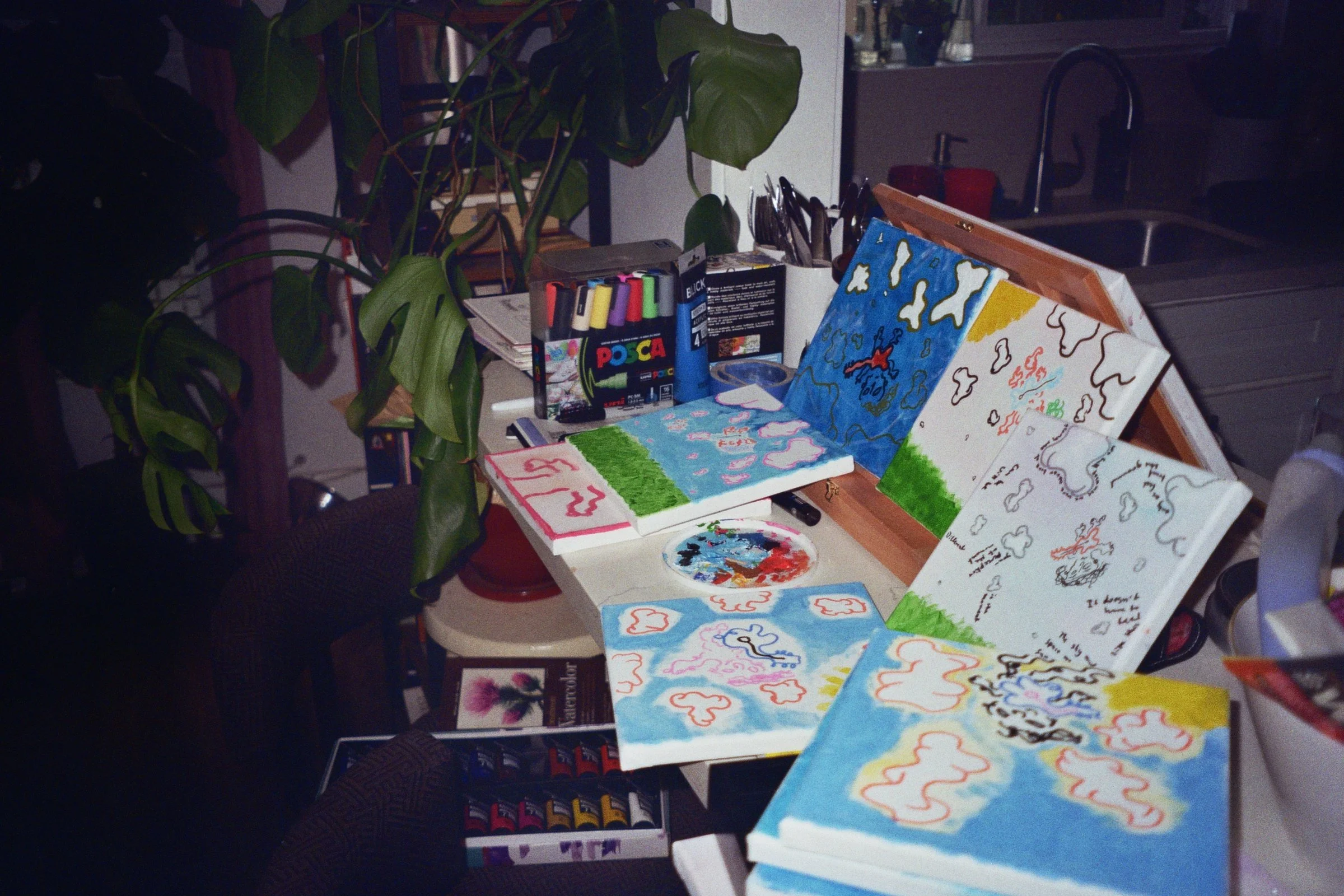 A cluttered desk with multiple small paintings and drawings of clouds, skies, and nature scenes in progress, surrounded by markers, paint supplies, and plants.