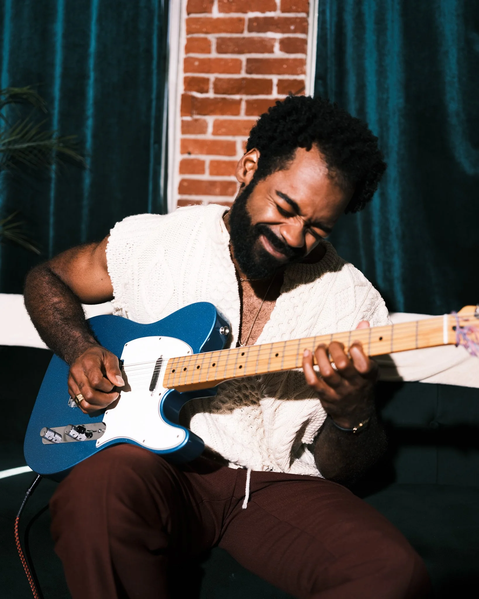 A man with a beard and curly hair playing an electric guitar, sitting indoors with a brick wall and teal curtains in the background.