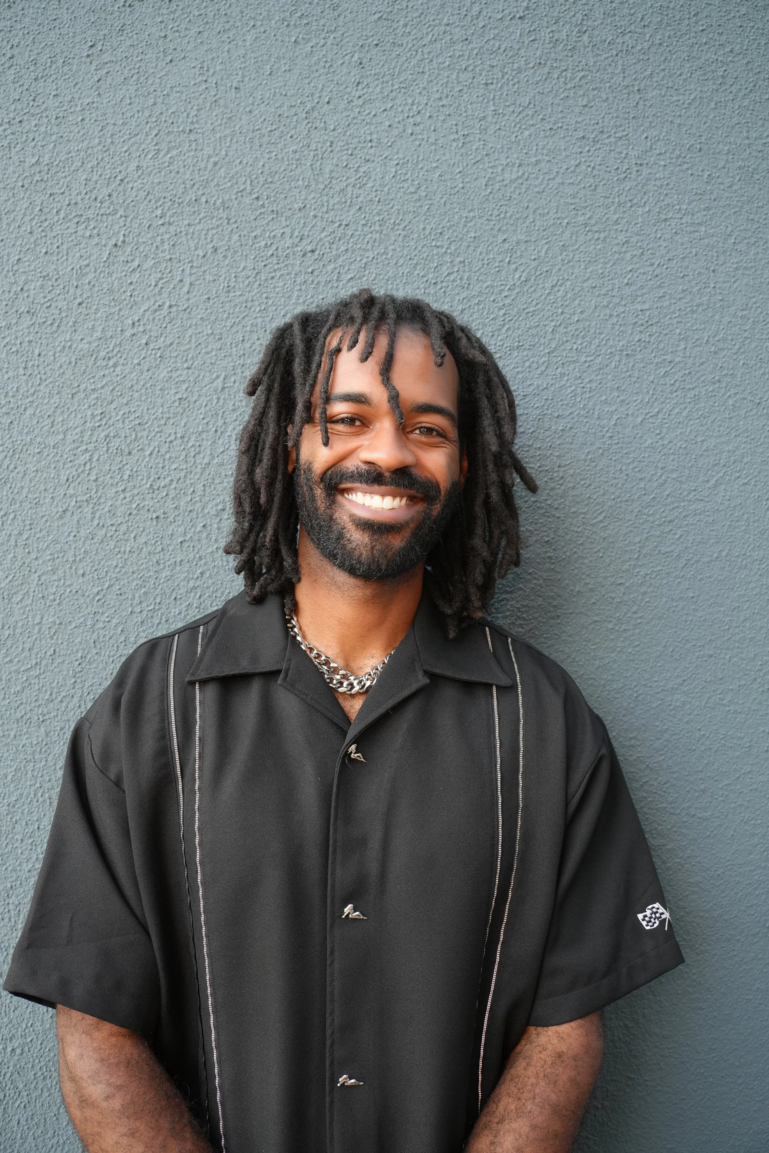 A man with dreadlocks, a beard, and a big smile standing against a textured gray wall, wearing a black short-sleeved shirt and a silver chain necklace.