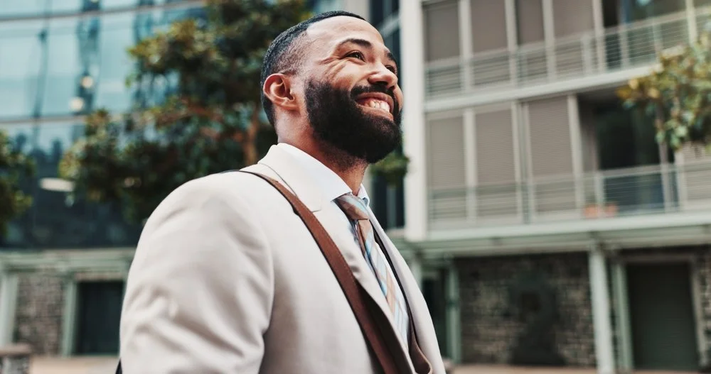 A smiling man in a suit stands outdoors with modern buildings and trees in the background.