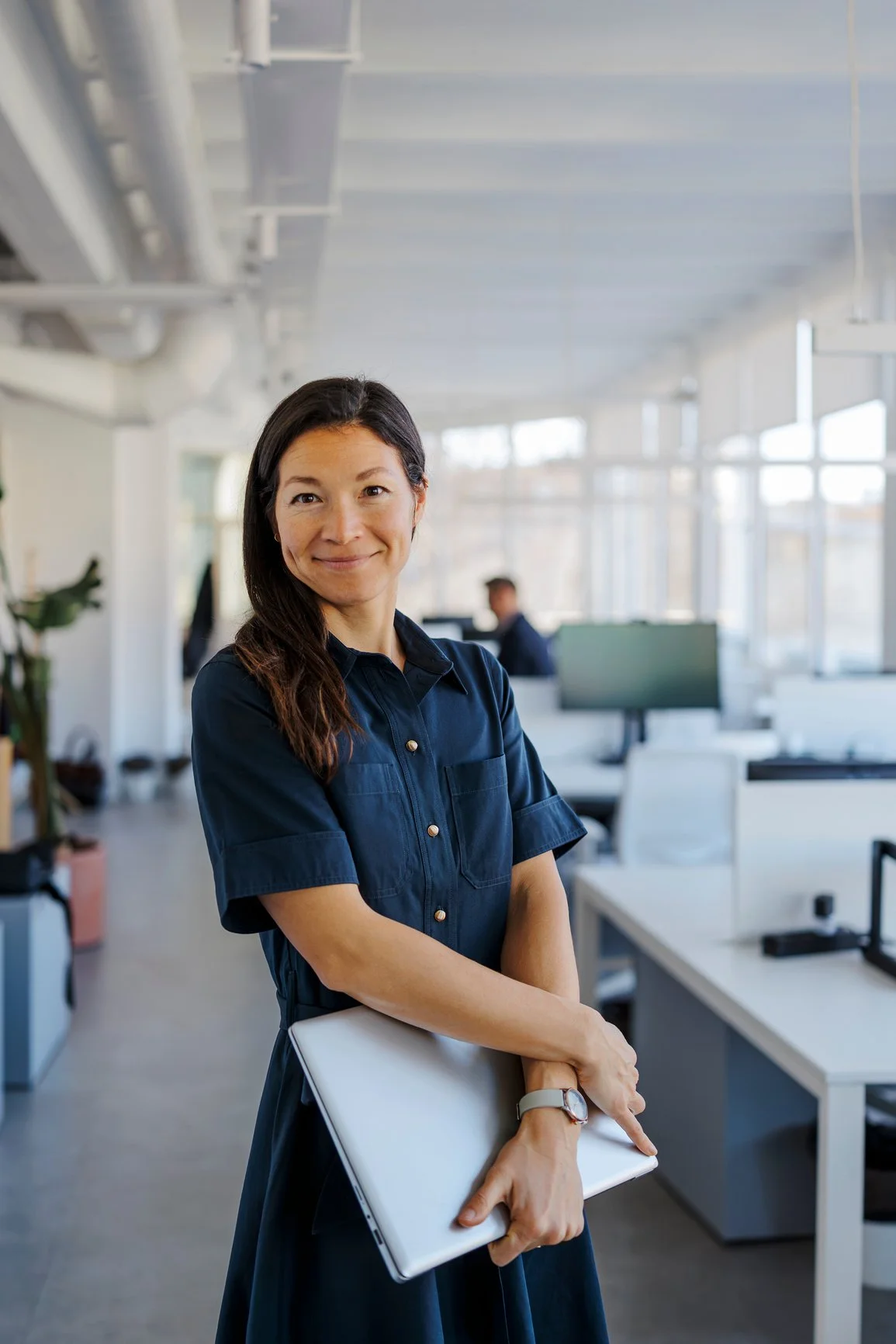 A woman standing in an office, smiling, holding a closed laptop, wearing a dark blue dress, with office desks and other people working in the background.