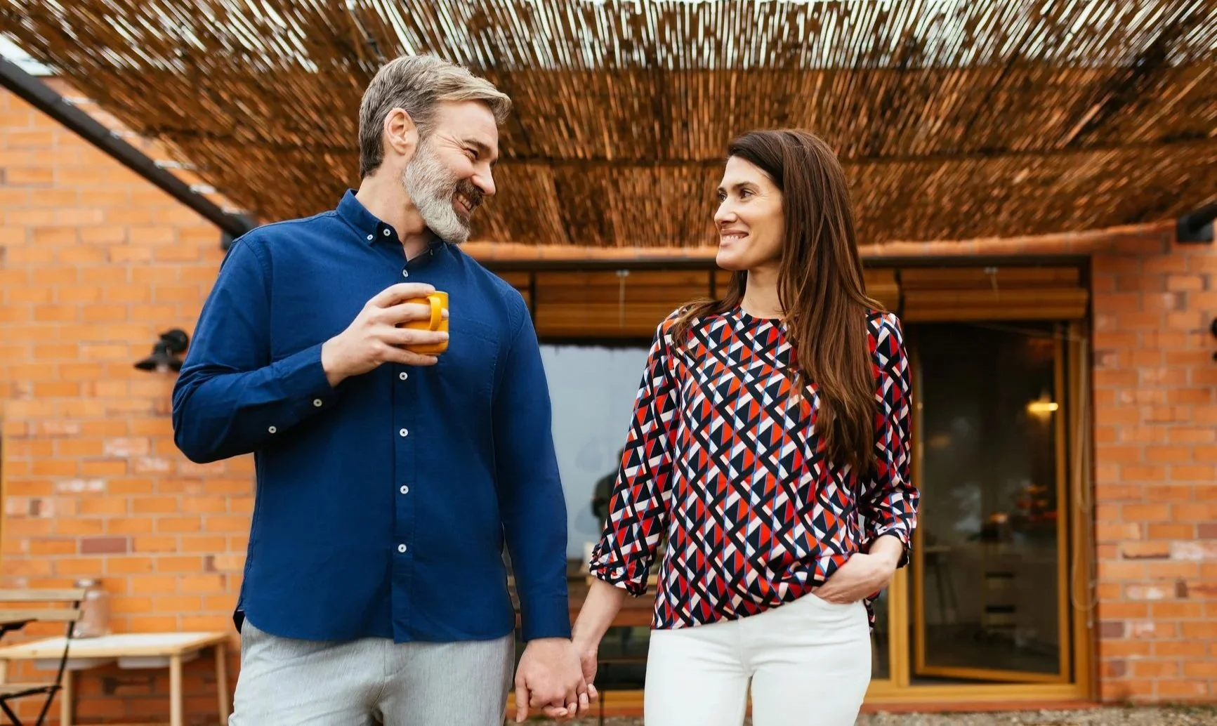 A middle-aged man with a beard holding a yellow mug and smiling at a woman with long brown hair, wearing a patterned blouse, who is smiling back. They are standing outside in front of a brick house with a wooden cover overhead.