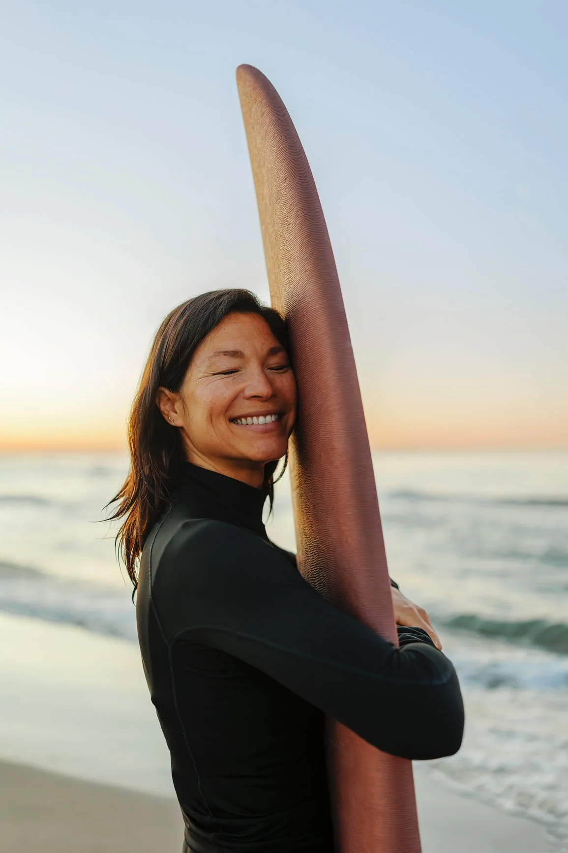 Woman smiling with eyes closed, holding a red surfboard on the beach at sunset.