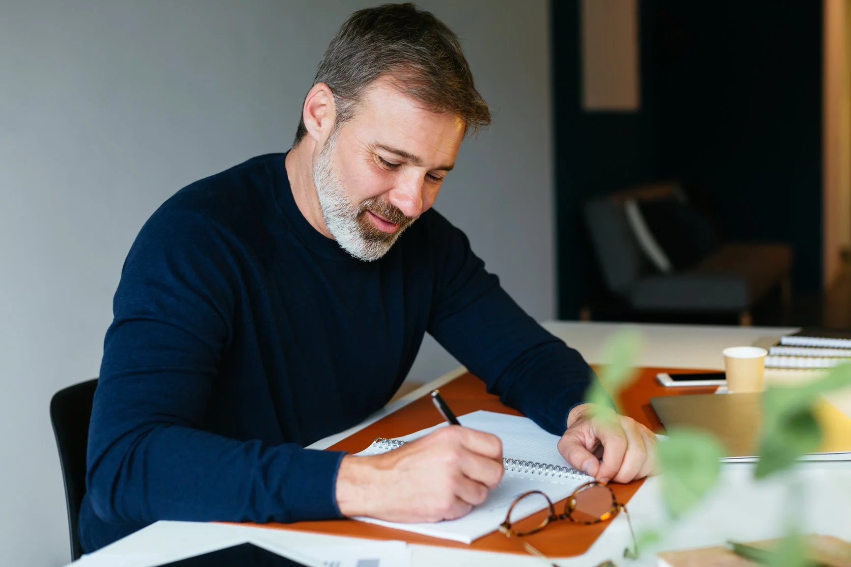 A middle-aged man with gray hair and a beard, wearing a dark blue shirt, is sitting at a desk writing in a notebook with a pen.