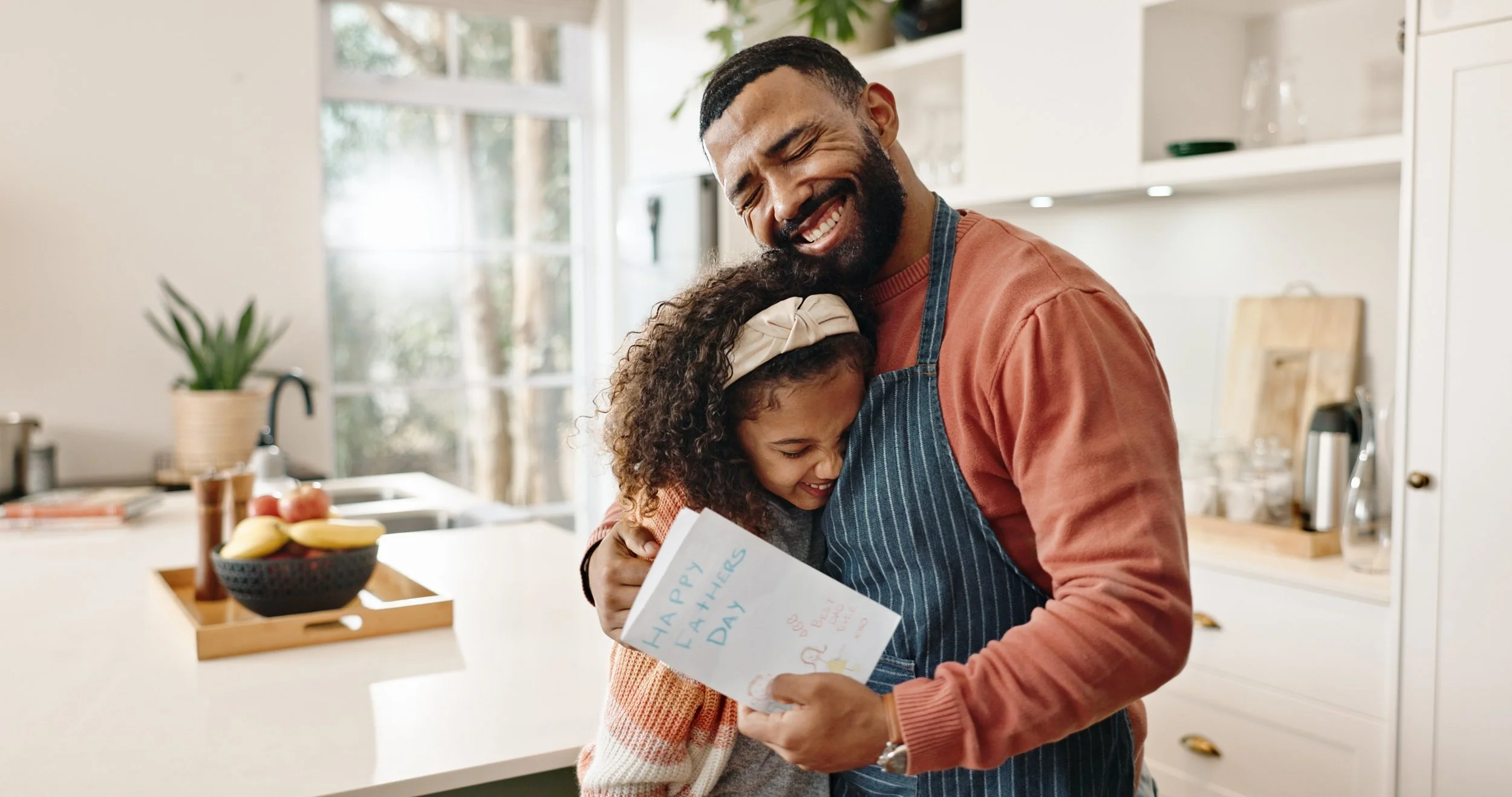 A man and young girl hugging in a bright kitchen, with the girl holding a birthday card that says "Happy Happy Friends Day," surrounded by a bowl of fruit, plants, and kitchen appliances.