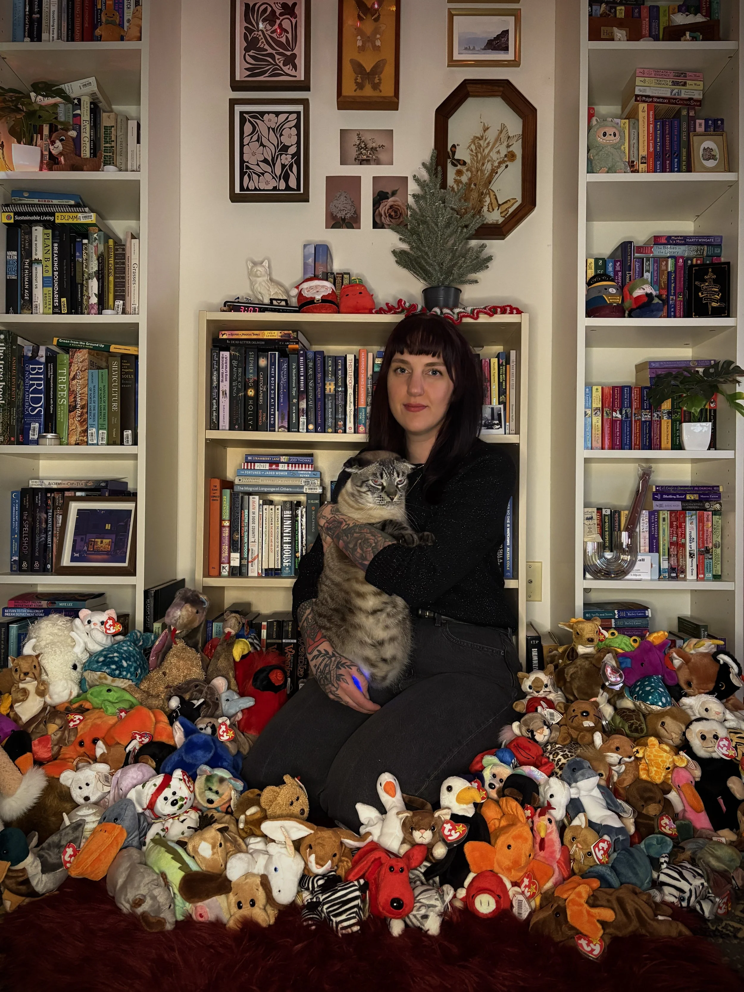 A woman with dark hair and tattoos on her arm kneeling on the floor and holding a tabby cat in a room decorated for Christmas with numerous plush toys surrounding her. There are bookshelves in the background filled with books and framed artwork and photographs on the wall.