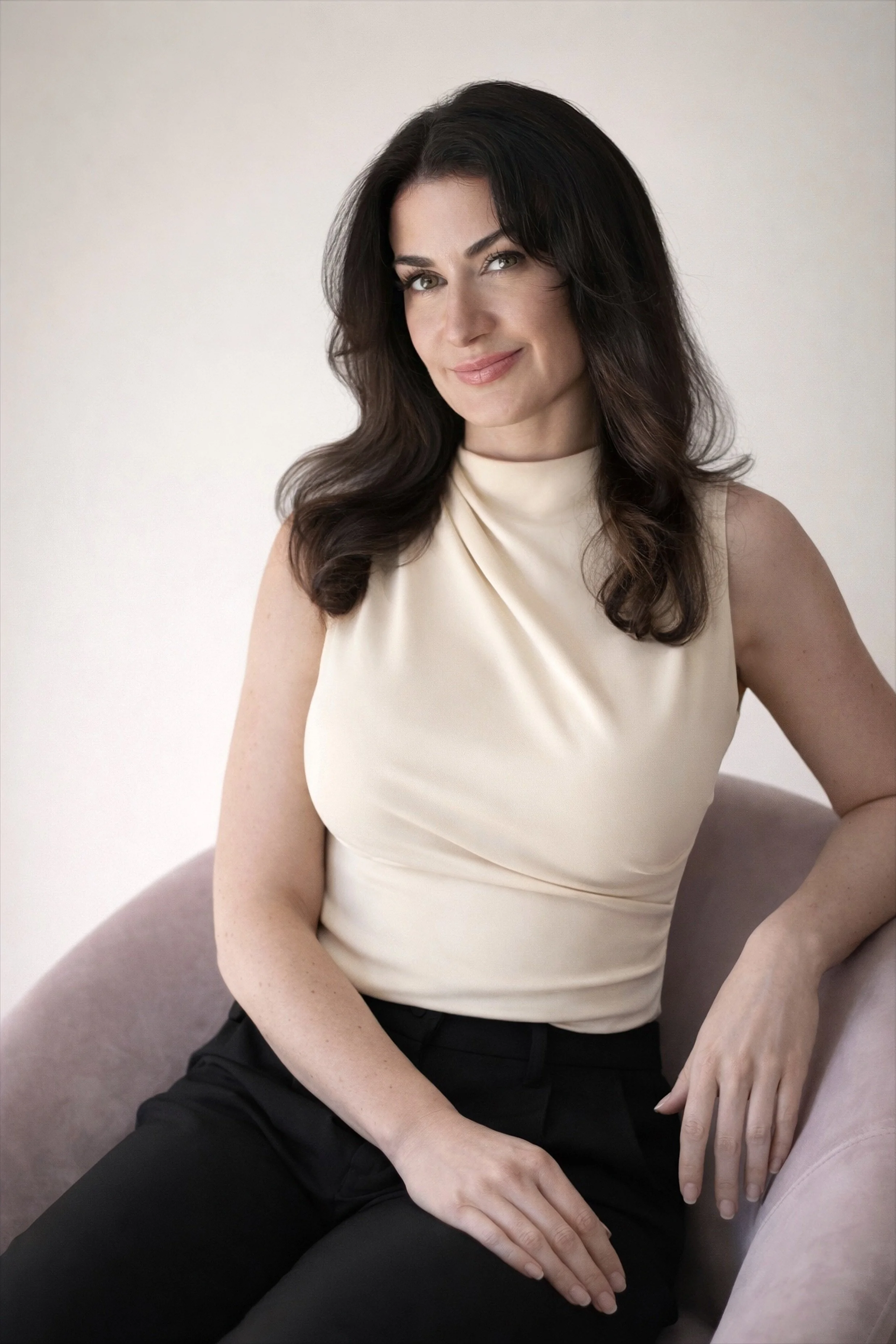 A woman with dark, wavy hair and fair skin smiling while sitting on a light-colored chair, wearing a sleeveless cream-colored top and black pants.