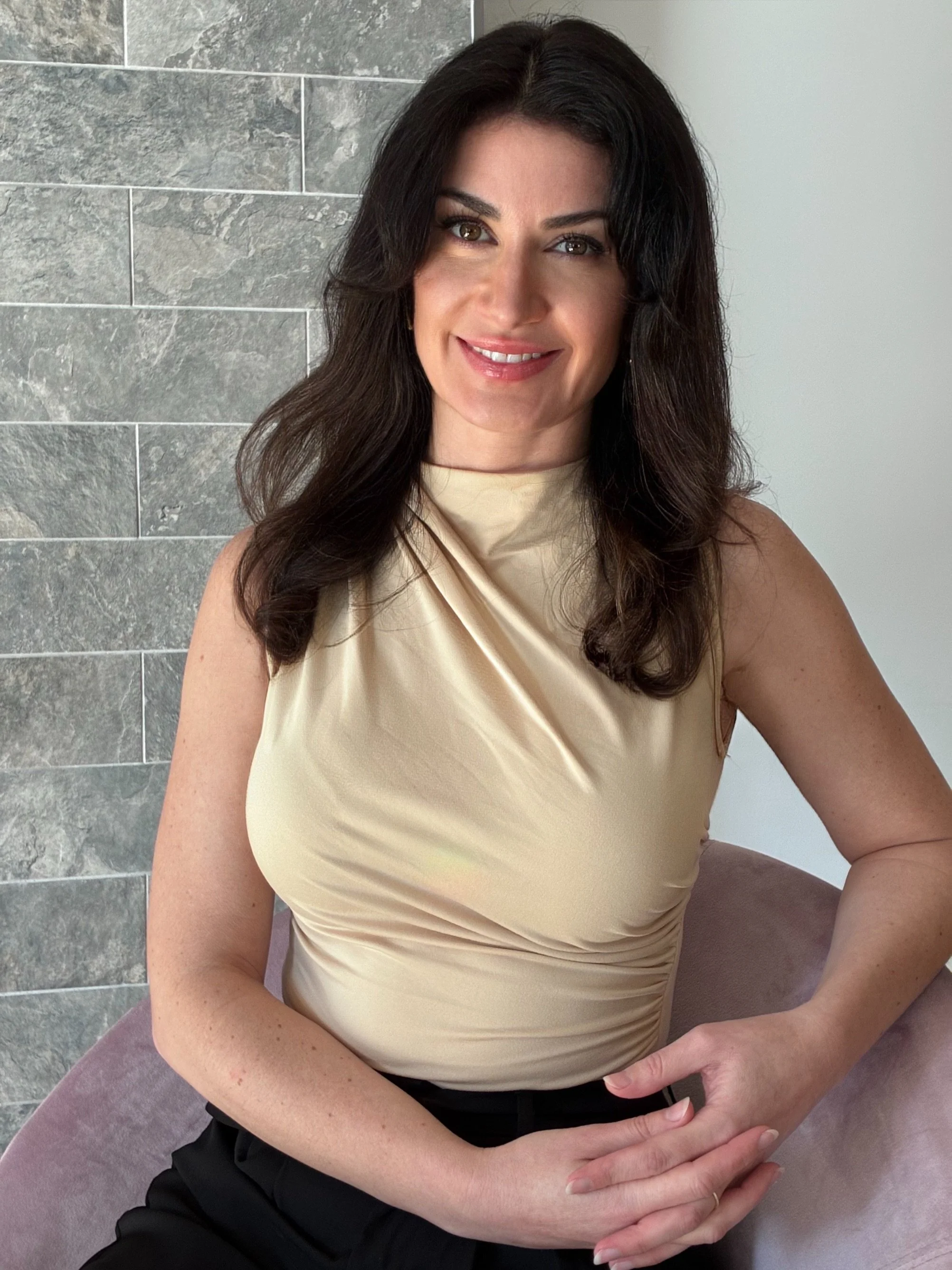 A woman with long dark hair, wearing a beige sleeveless top and black pants, sitting on a purple chair with a gray stone wall behind her, smiling at the camera.