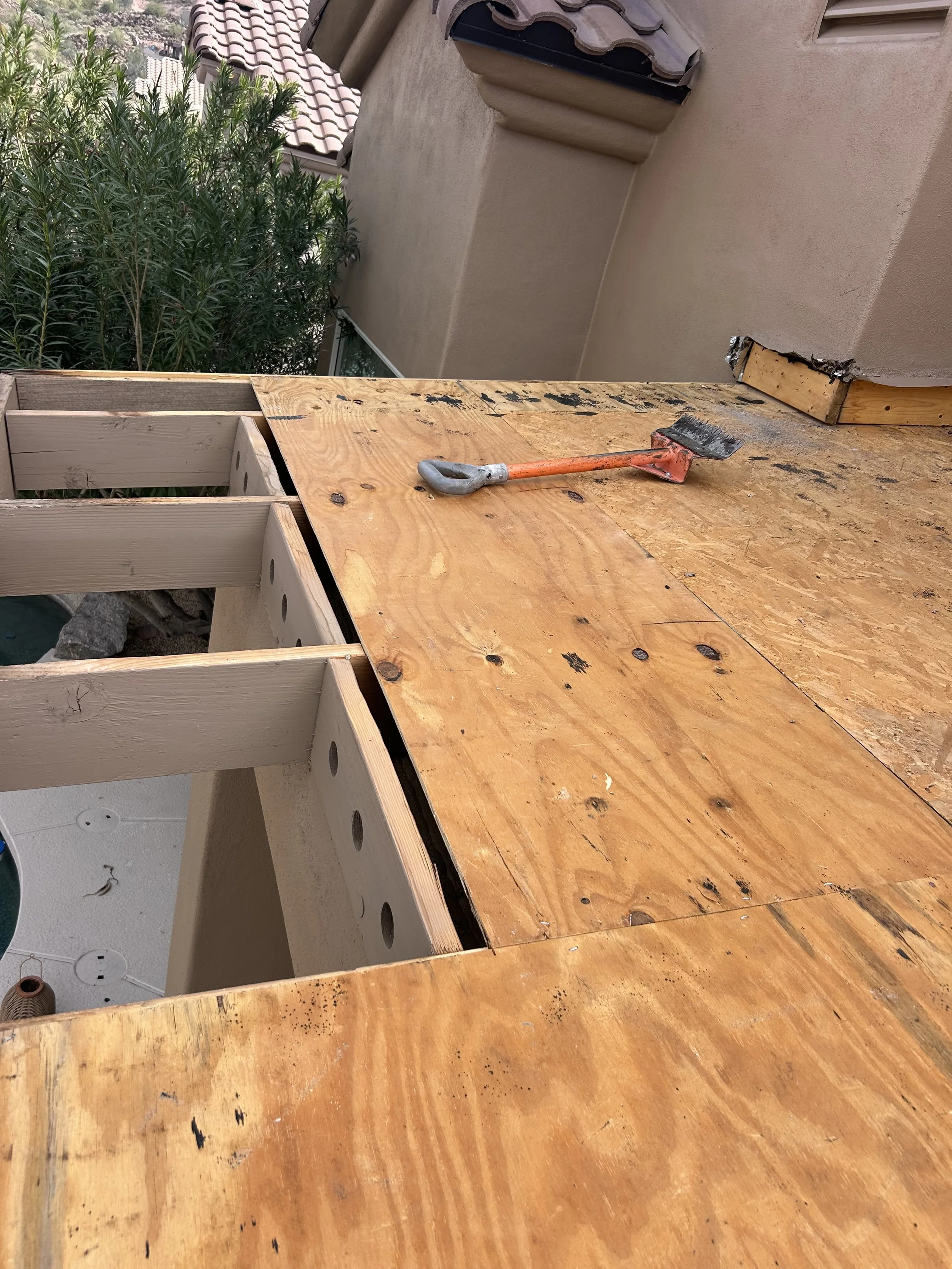 A partially built outdoor deck shows plywood sheets on top of wooden framing, with a small black and orange shovel resting on the surface.