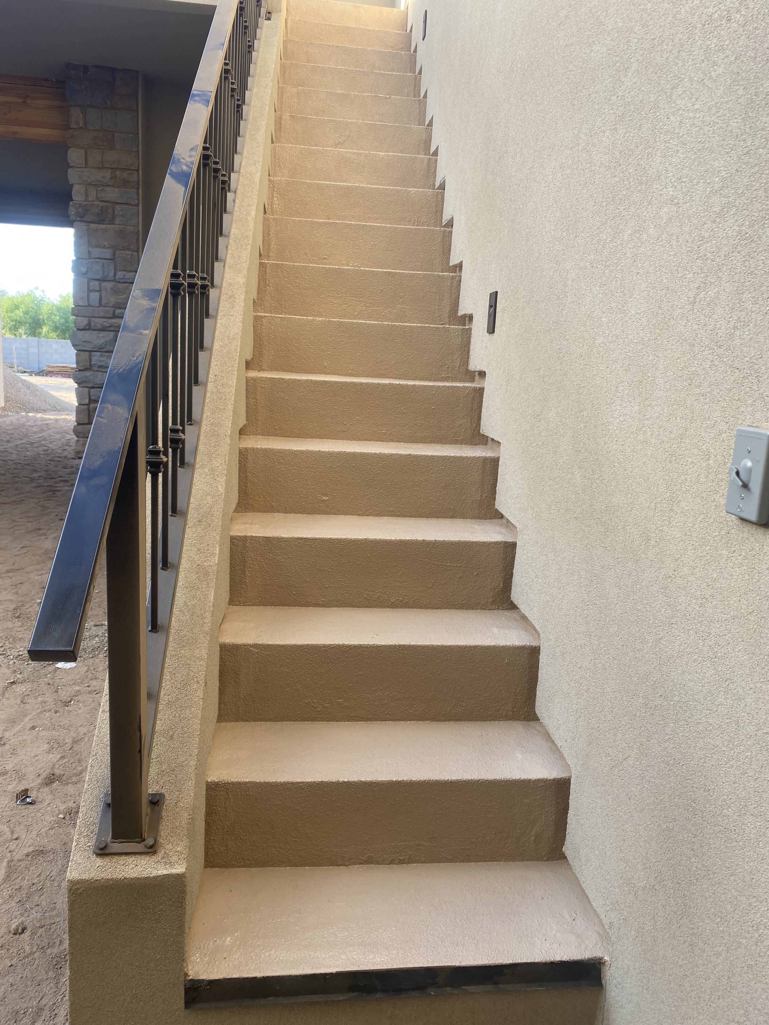 Exterior beige staircase with black metal railing on the left side, next to a cream-colored wall with a light switch, leading upwards. Part of a stone column and window are visible on the left.