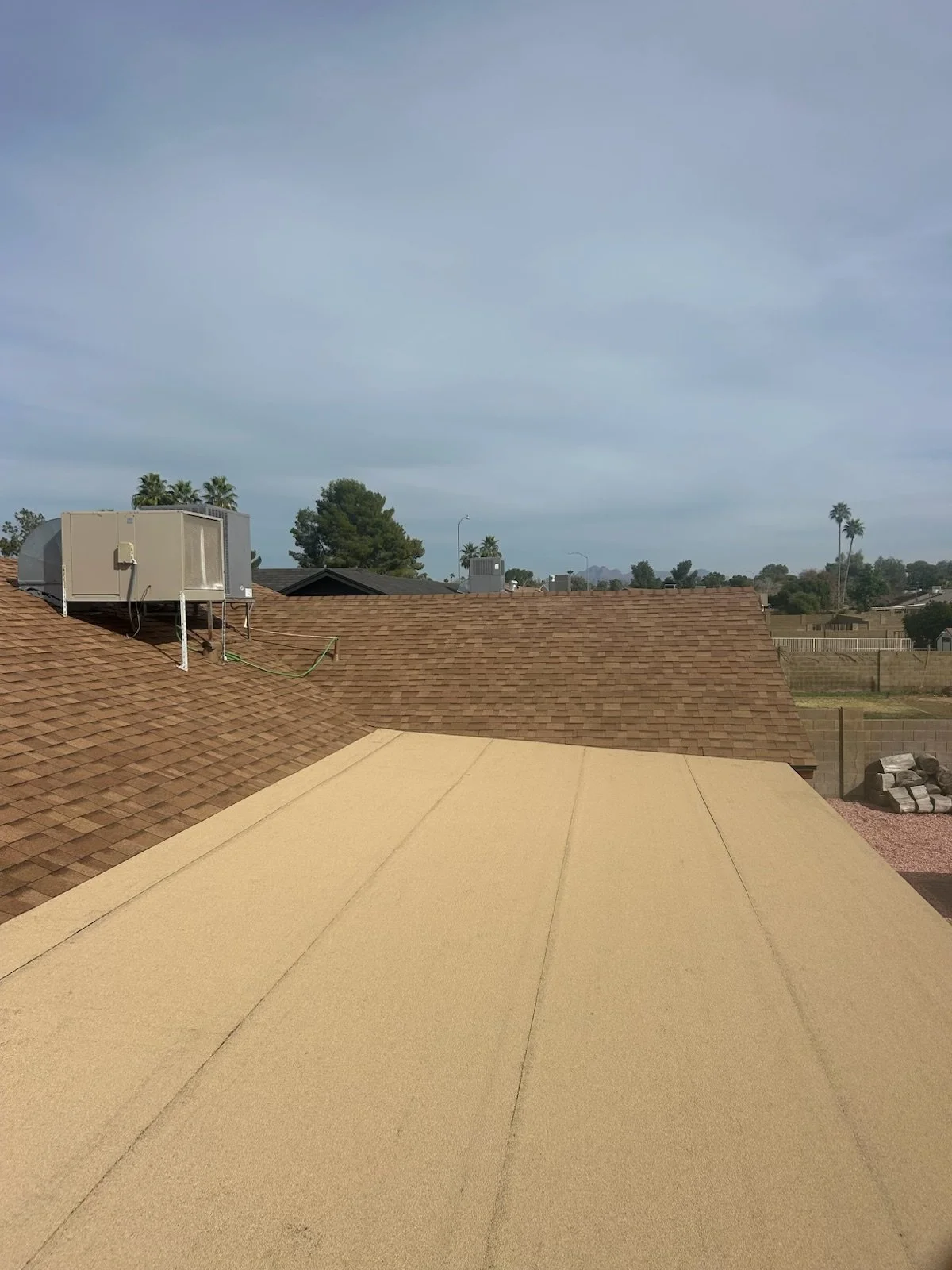 View of rooftops with brown shingles and a beige flat roof, with trees and a cloudy sky in the background.
