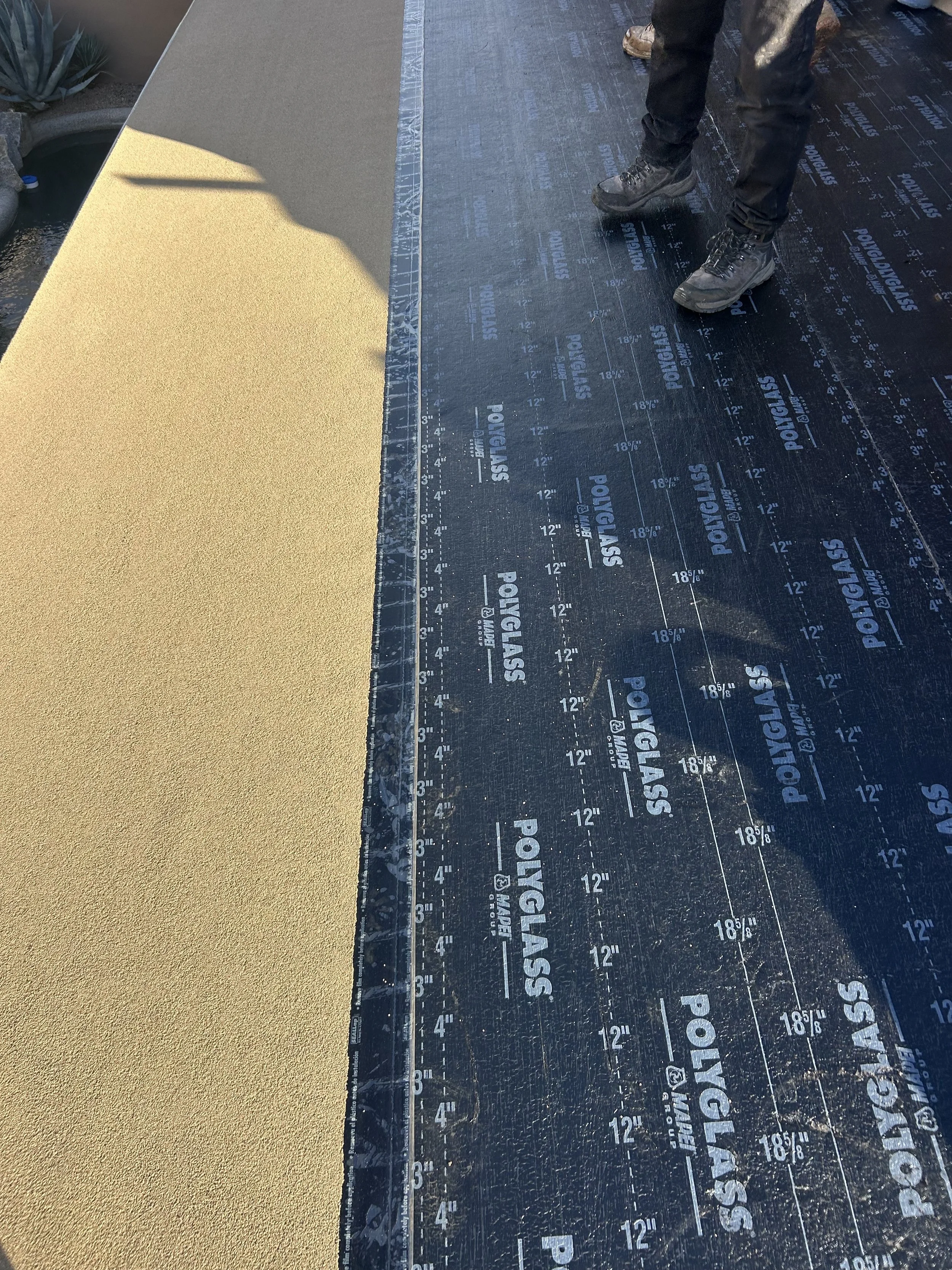 A construction worker standing on a black Polyglass roofing underlayment, next to a rolled sheet of tan-colored roofing material.