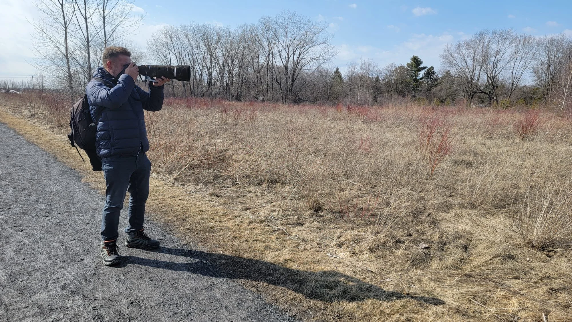 A man in a blue jacket, jeans, and hiking boots stands on a gravel path, taking a photograph with a long camera lens. He has a black backpack and is looking into the distance. There are leafless trees and dry grass in the background under a partly cloudy sky.