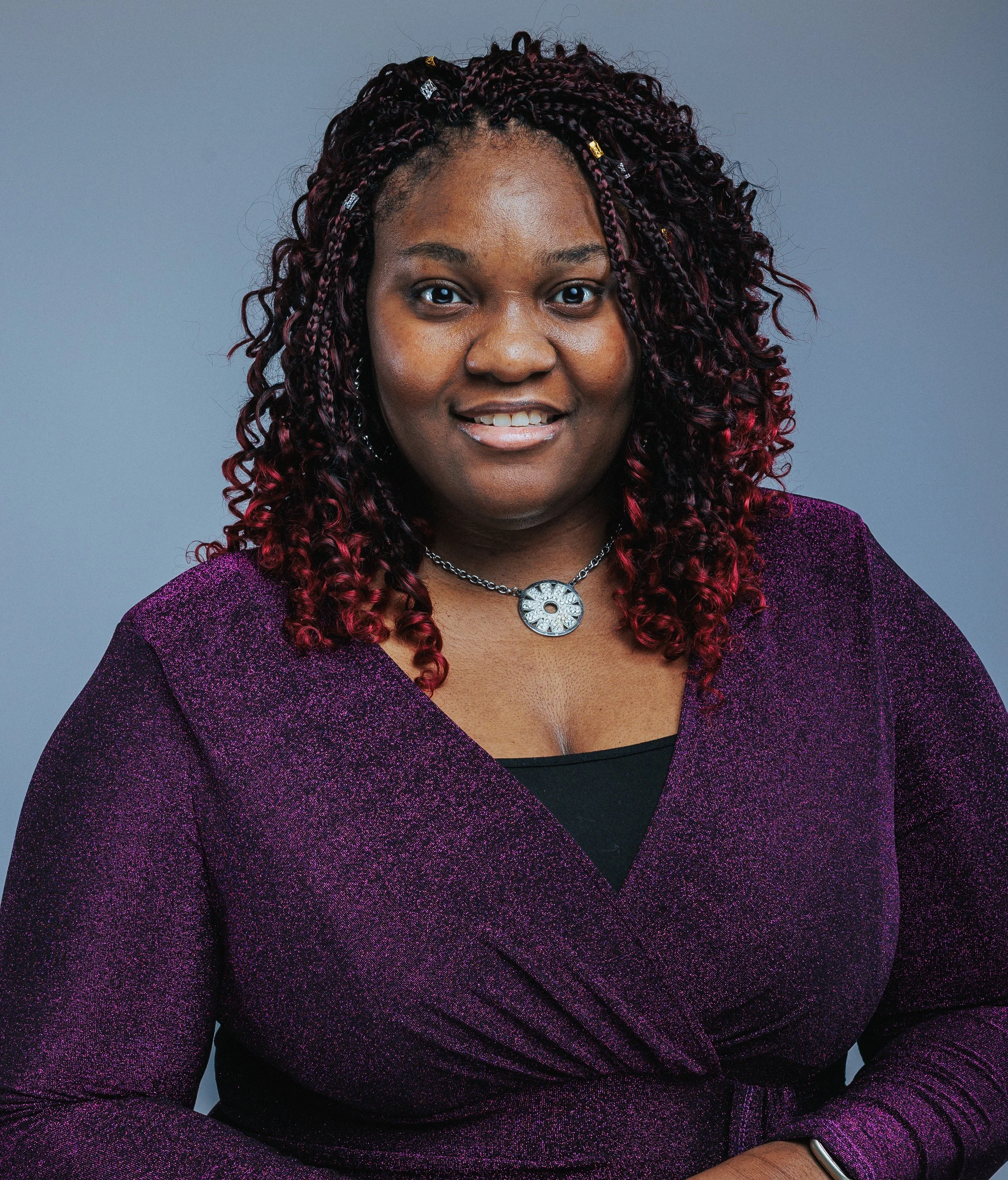 Portrait of a woman with dark skin, curly red hair, wearing a purple glittery top, a silver necklace, and a bracelet, against a gray background.