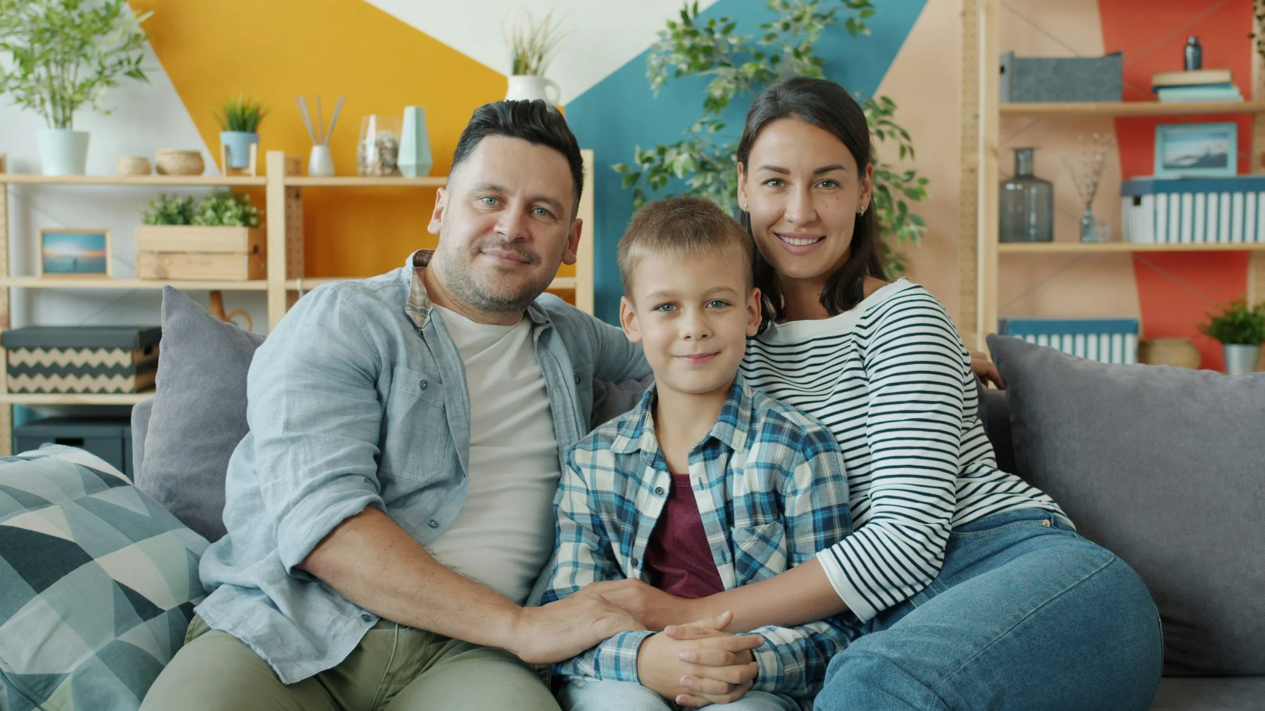 Family of three sitting on a couch in a colorful living room, smiling and looking at the camera.