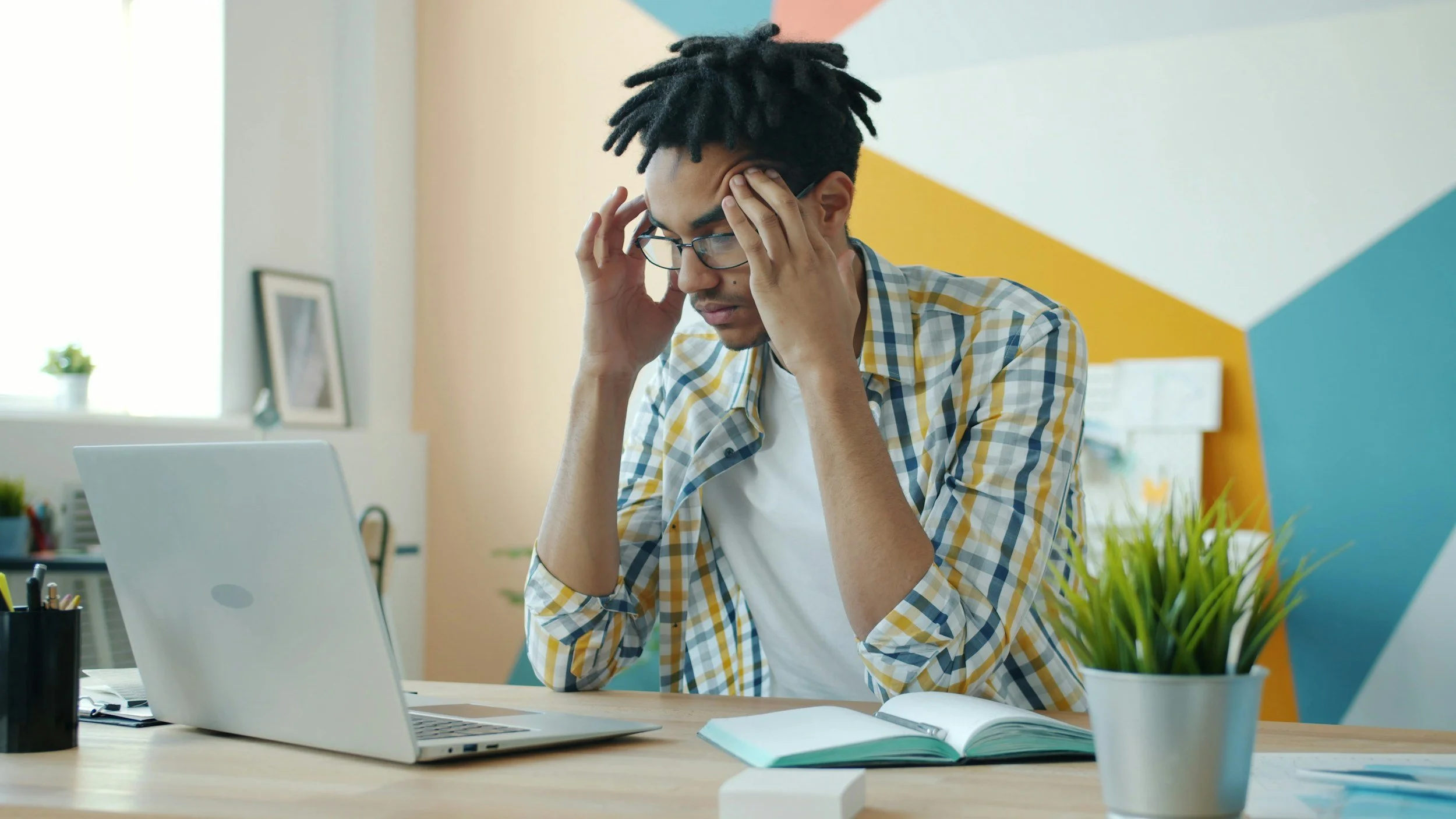 Young man with glasses, wearing a plaid shirt, sitting at a desk with a laptop, holding his head in frustration, with an open book and a potted plant on the desk.