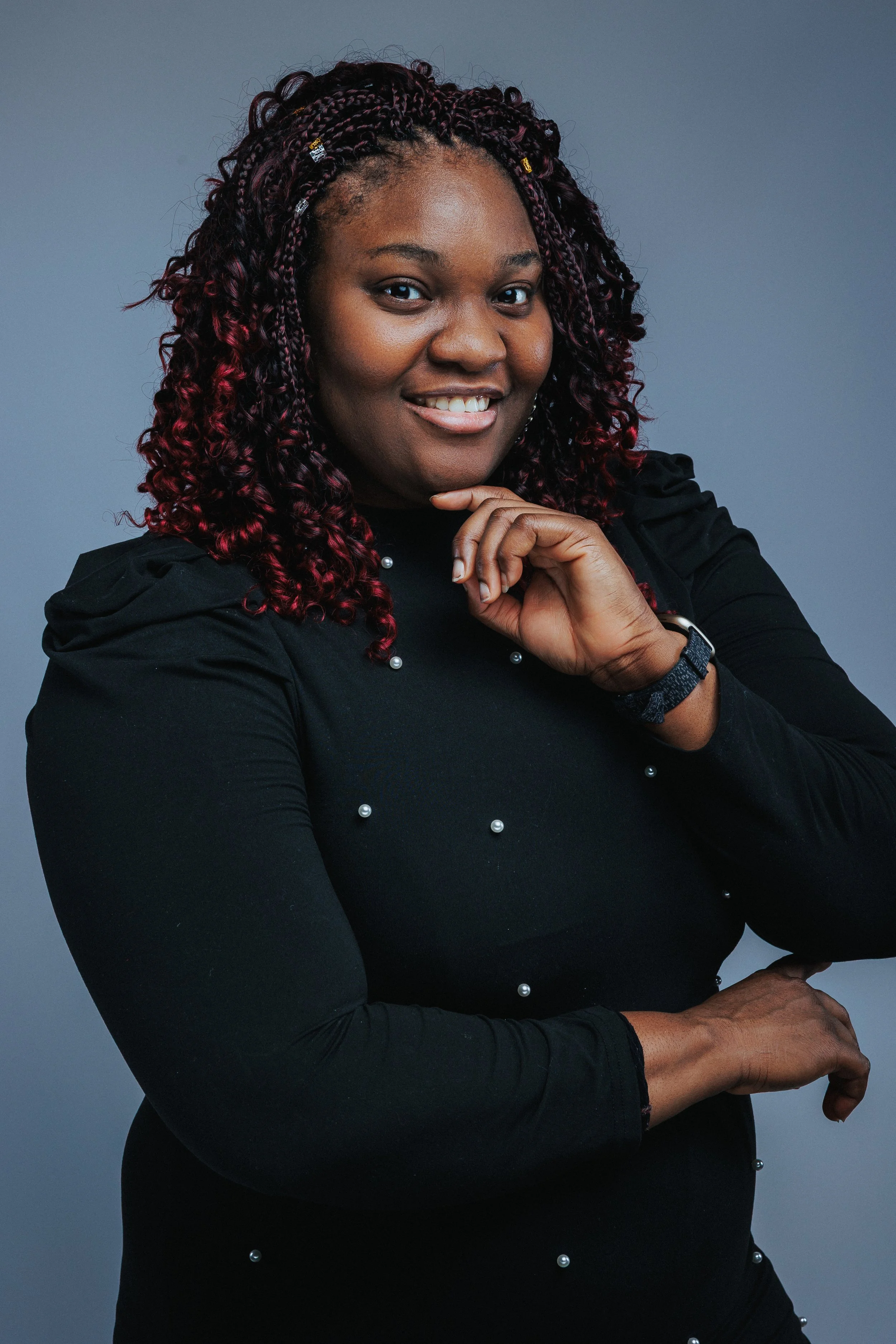 A woman with dark skin and curly hair with red highlights, wearing a black long-sleeve top with pearl embellishments, smiling and posing with her hand on her chin against a neutral gray background.